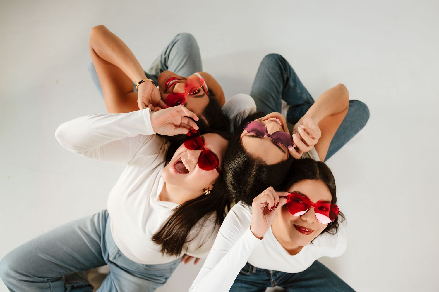 Three young women lying down wearing red sunglasses and casual outfits, laughing and posing together.