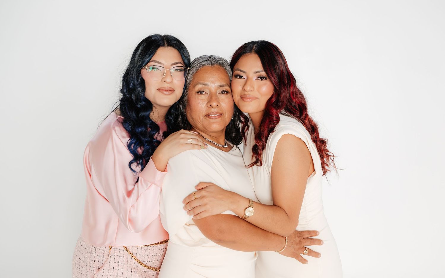 Three women pose together warmly against a white background, dressed in elegant pastel outfits for a family portrait.
