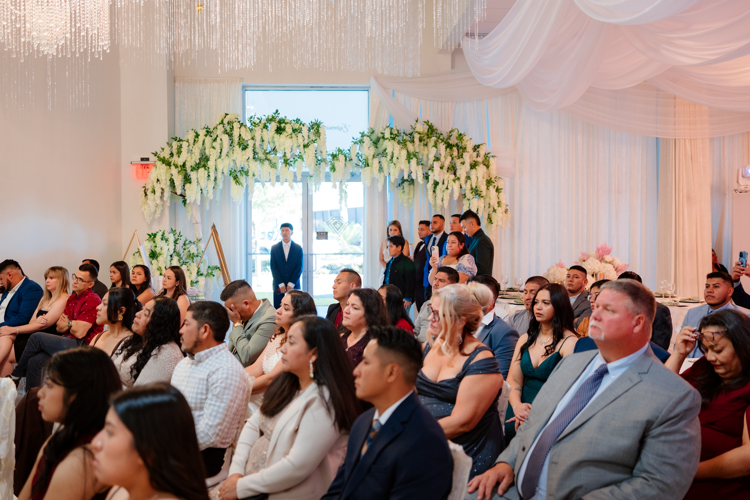 Guests seated at an elegant wedding ceremony with floral arch backdrop and white draped ceiling decor.