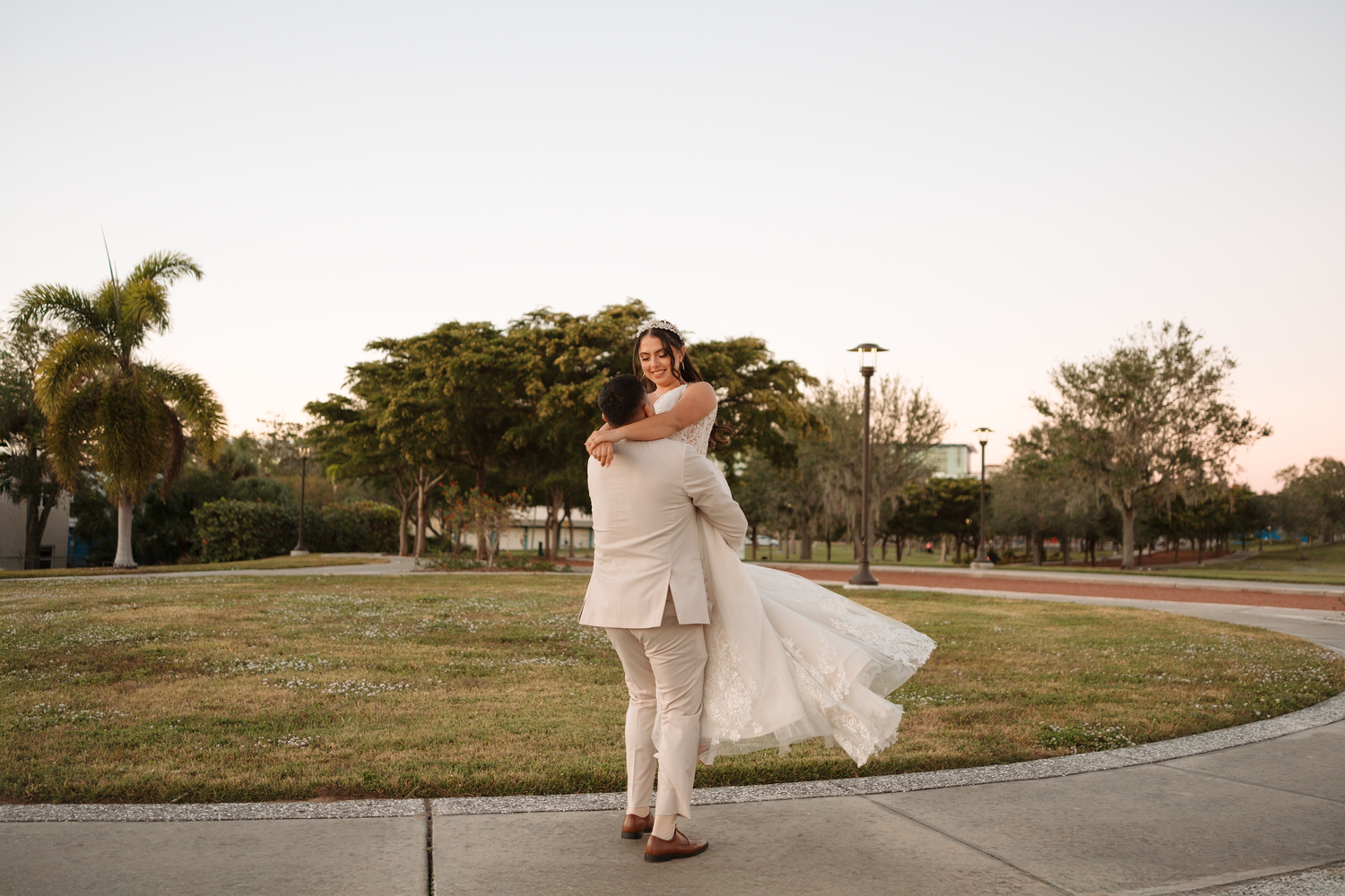 Groom lifting his bride in a white wedding dress outdoors at sunset with palm trees in the background.