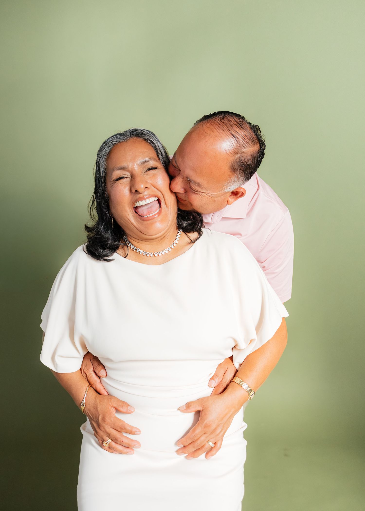 A joyful pregnant woman in white dress laughs as her partner kisses her cheek against a green background.