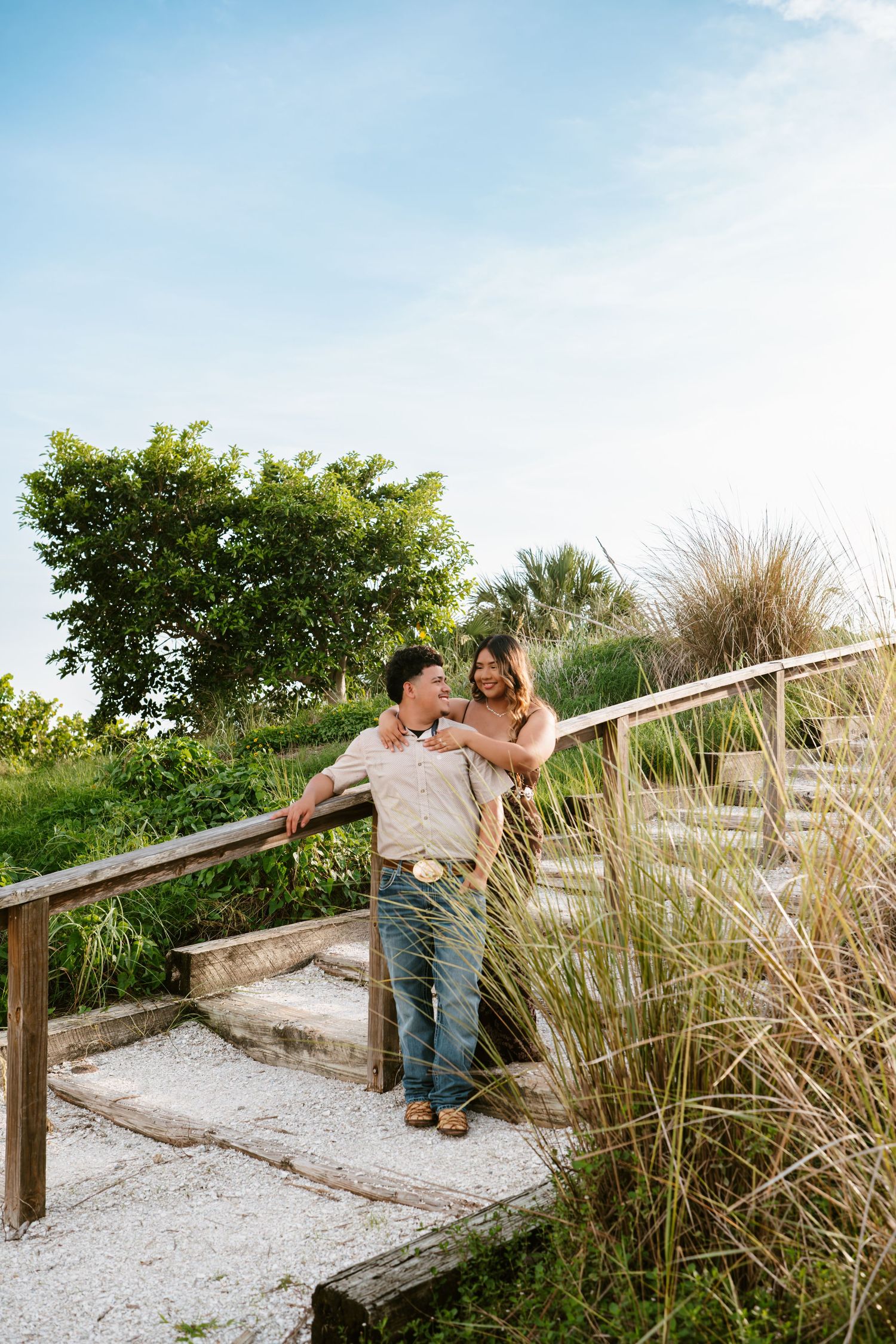 Couple embracing on wooden beach boardwalk steps surrounded by lush green coastal vegetation under a blue sky.