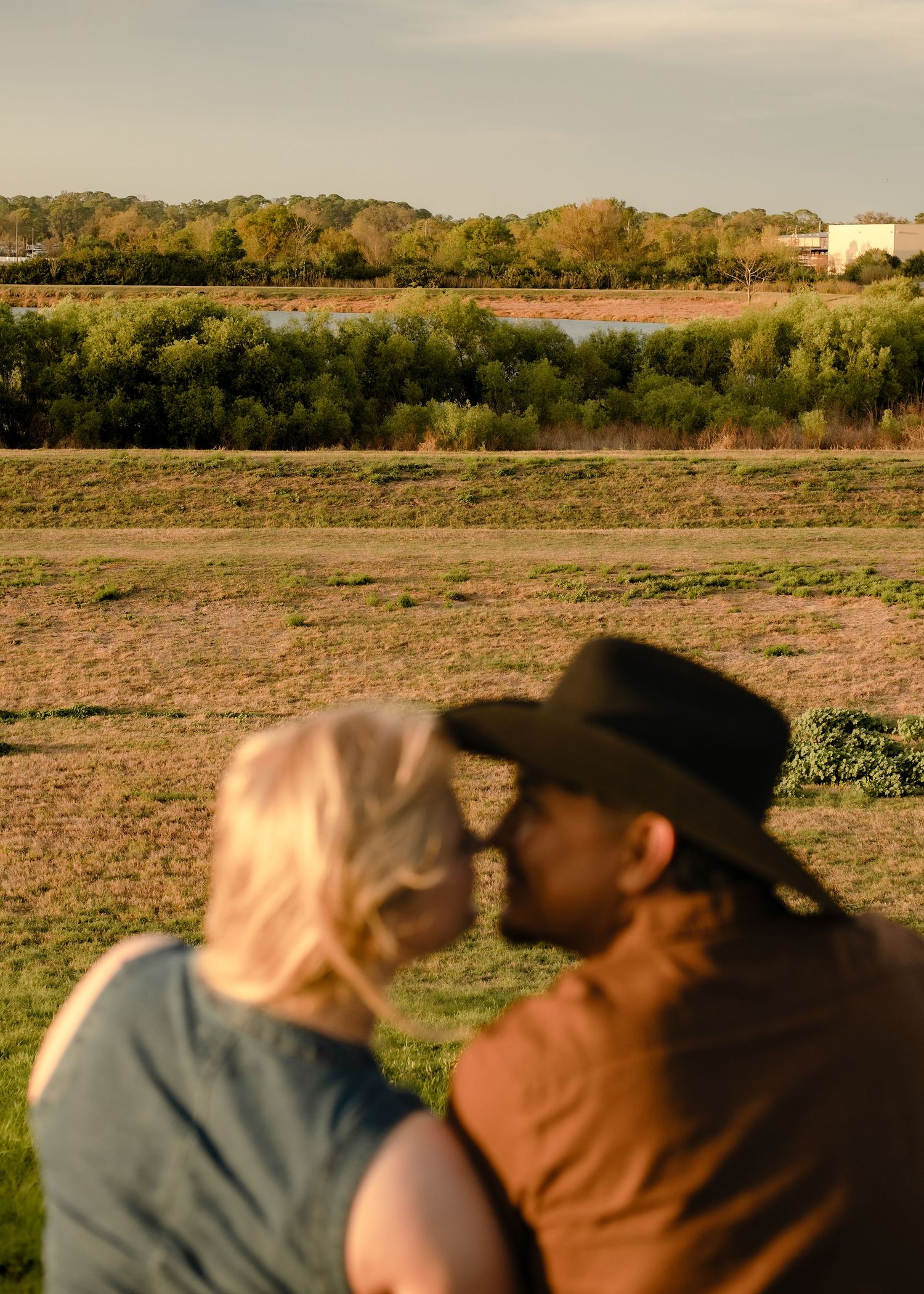 Couple sharing a romantic kiss outdoors near a river landscape at sunset, man wearing a cowboy hat.