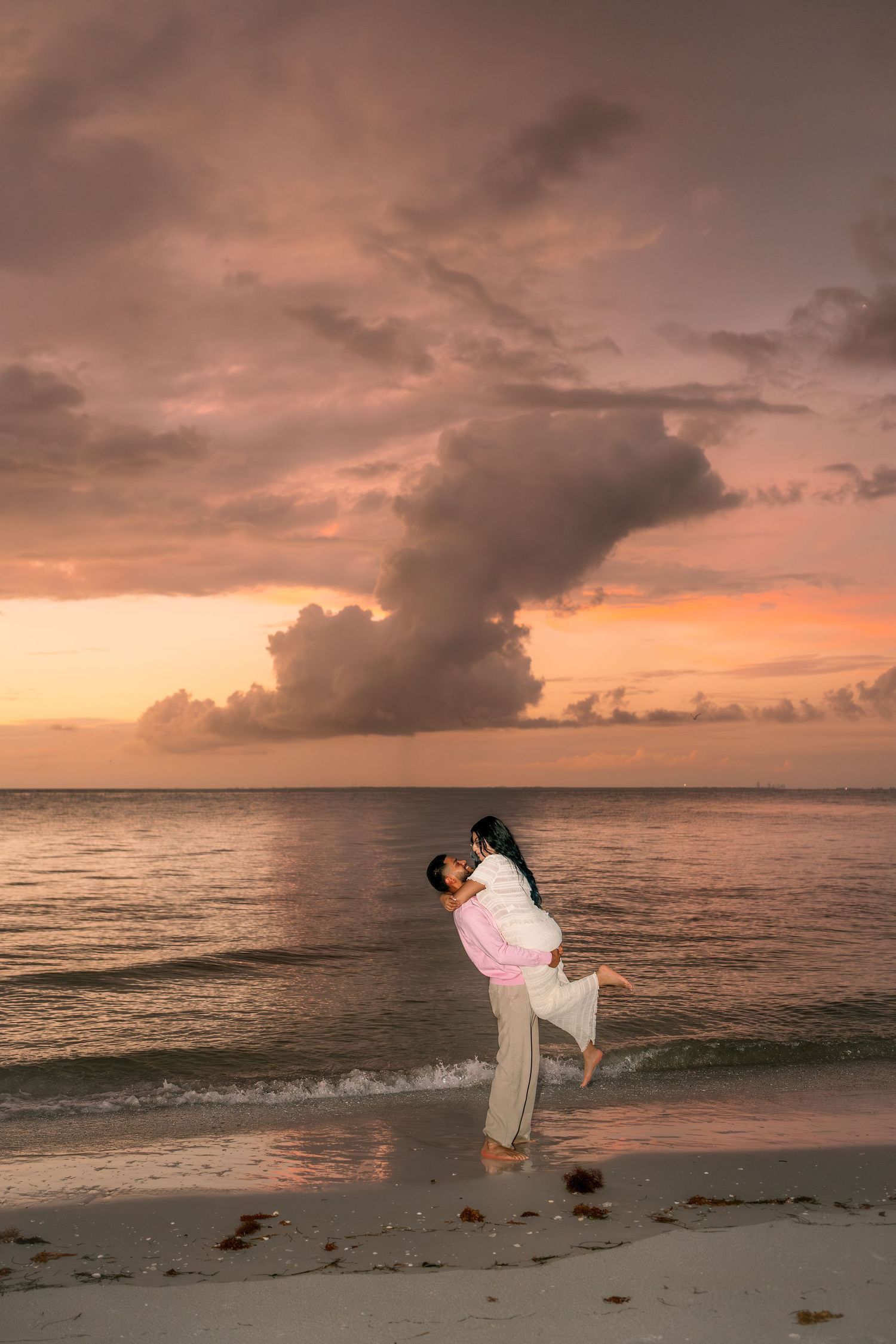 Couple embracing on a sandy beach at sunset with dramatic pink and orange clouds over the ocean.