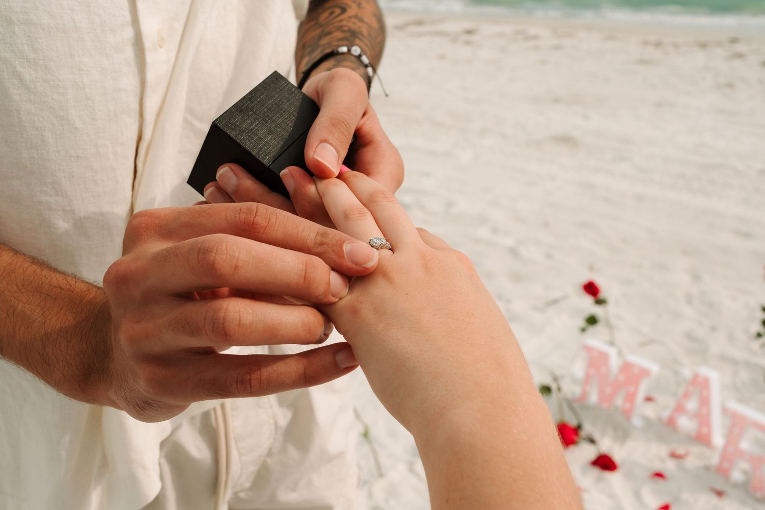 Man placing diamond engagement ring on woman's finger during romantic beach proposal with rose petals and decorative letters.