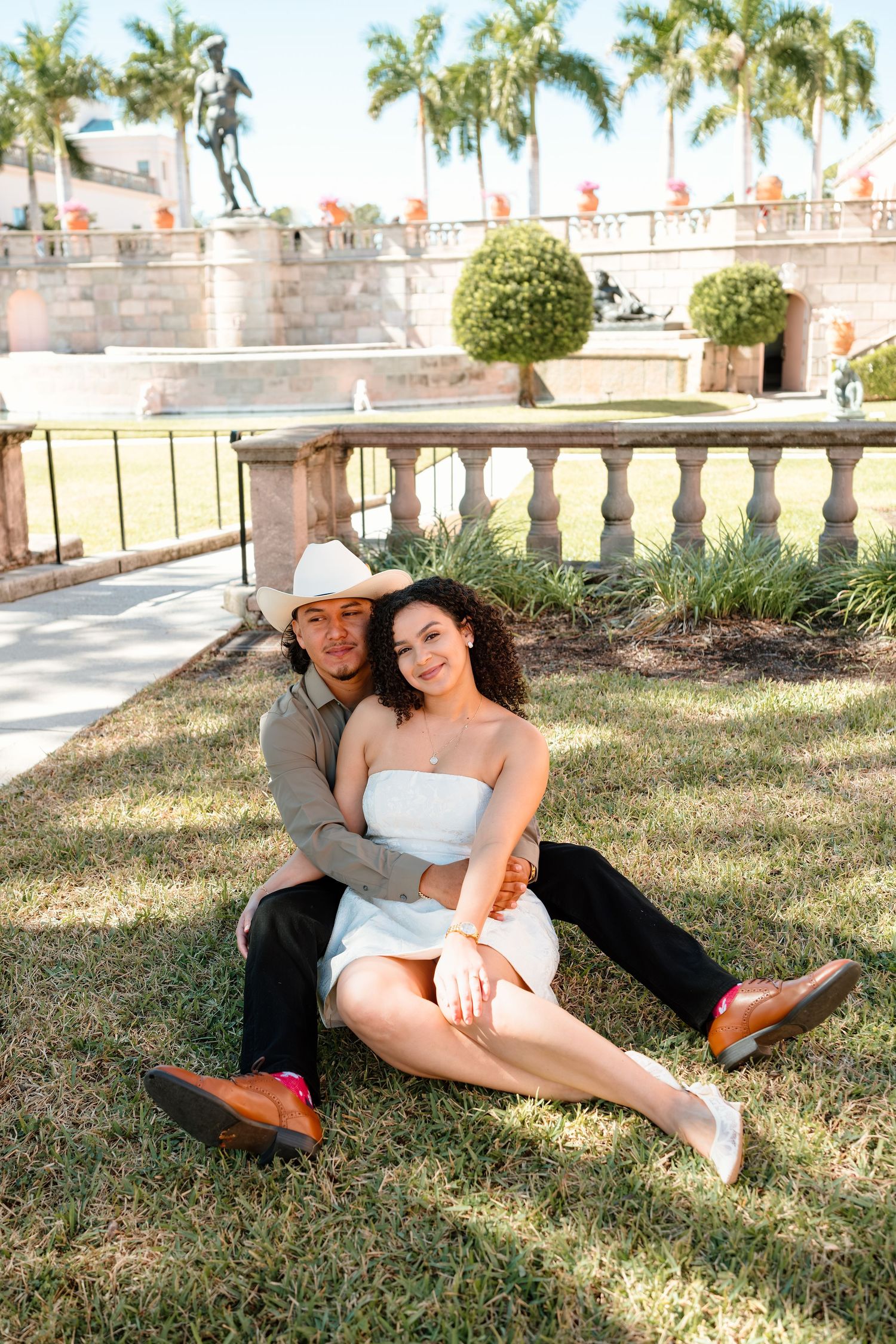 Couple sitting on grass in front of a fountain, woman in white dress and man in cowboy hat.
