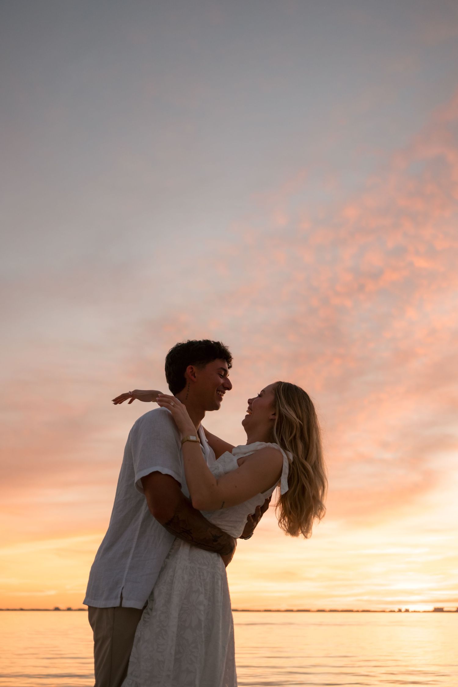 Romantic couple embracing at sunset by the water, silhouetted against a vibrant pink and orange sky.