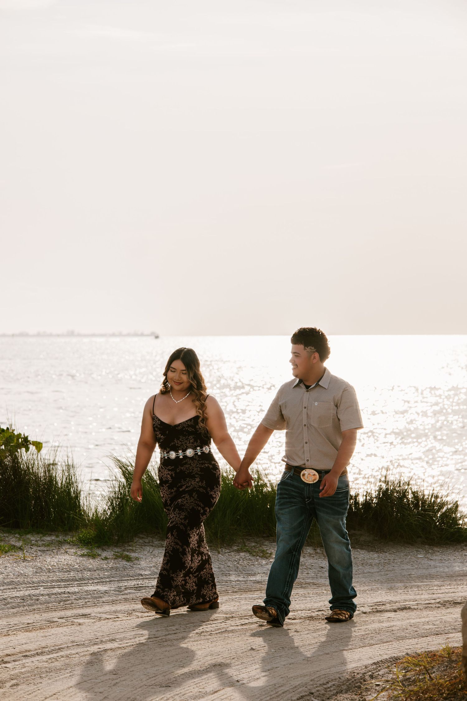 A couple holds hands and walks along a sandy path by the ocean at sunset, woman in black dress, man in jeans.