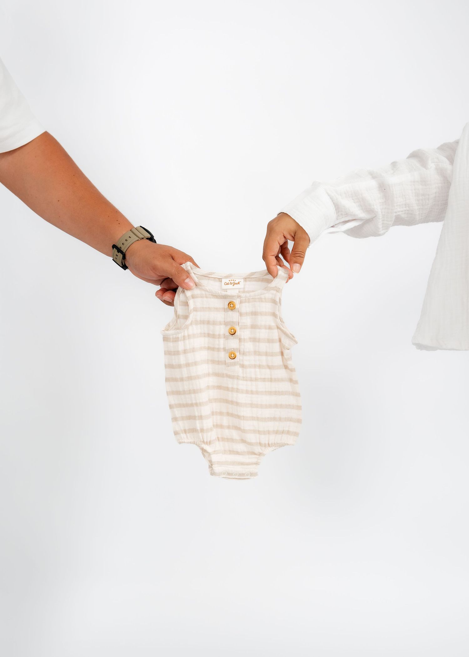 Two people holding a beige striped baby onesie with gold buttons against a white background.