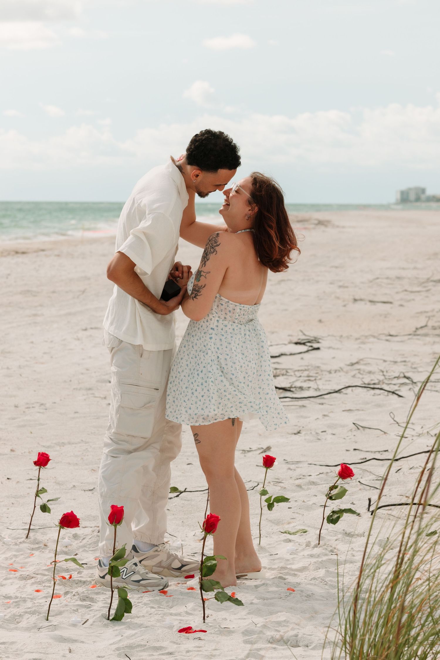 Romantic couple sharing a tender moment on a sandy beach surrounded by red roses with ocean views in the background.
