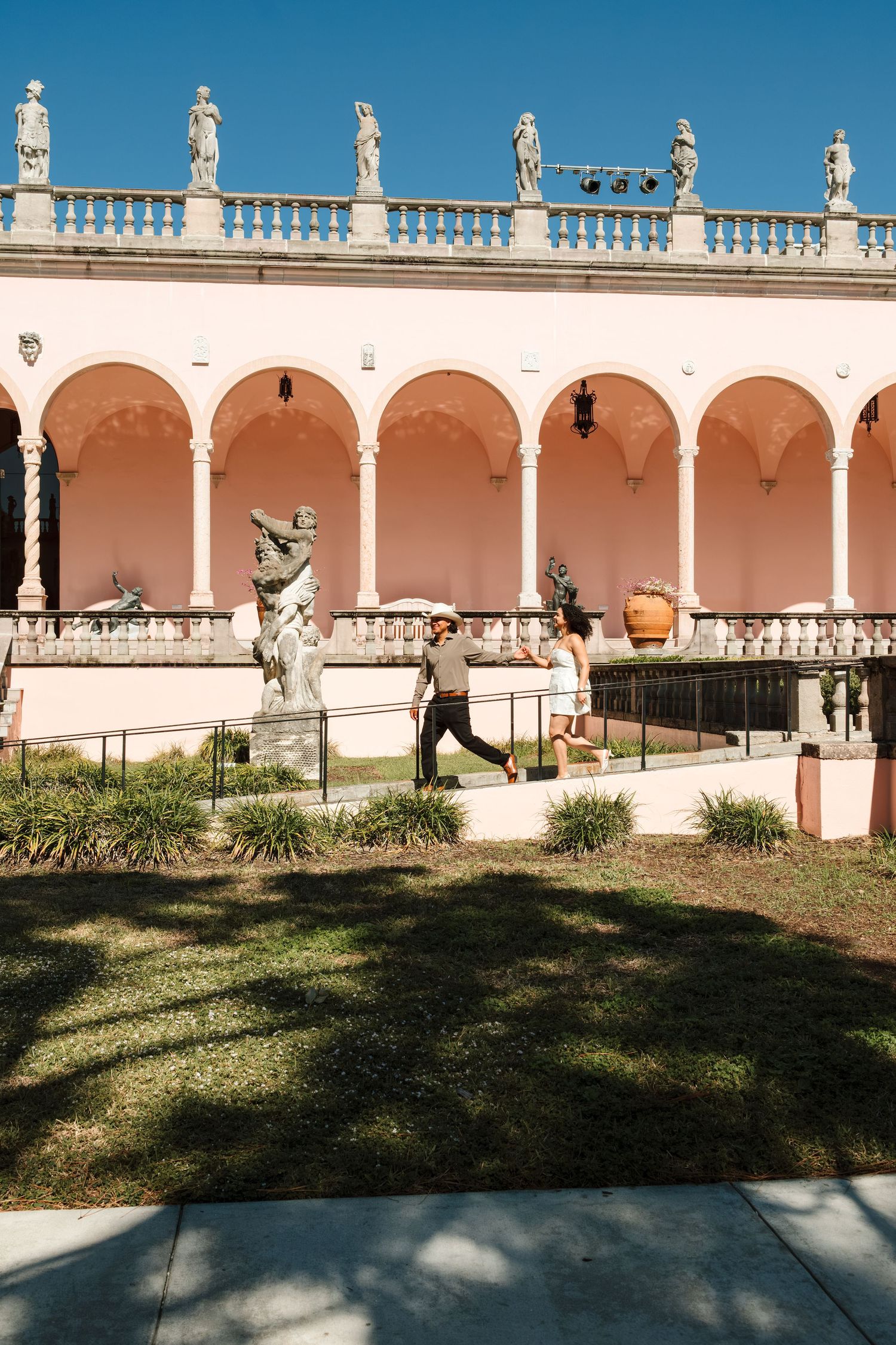 Couple walking through a pink Italian Renaissance-style courtyard with arches and classical sculptures.
