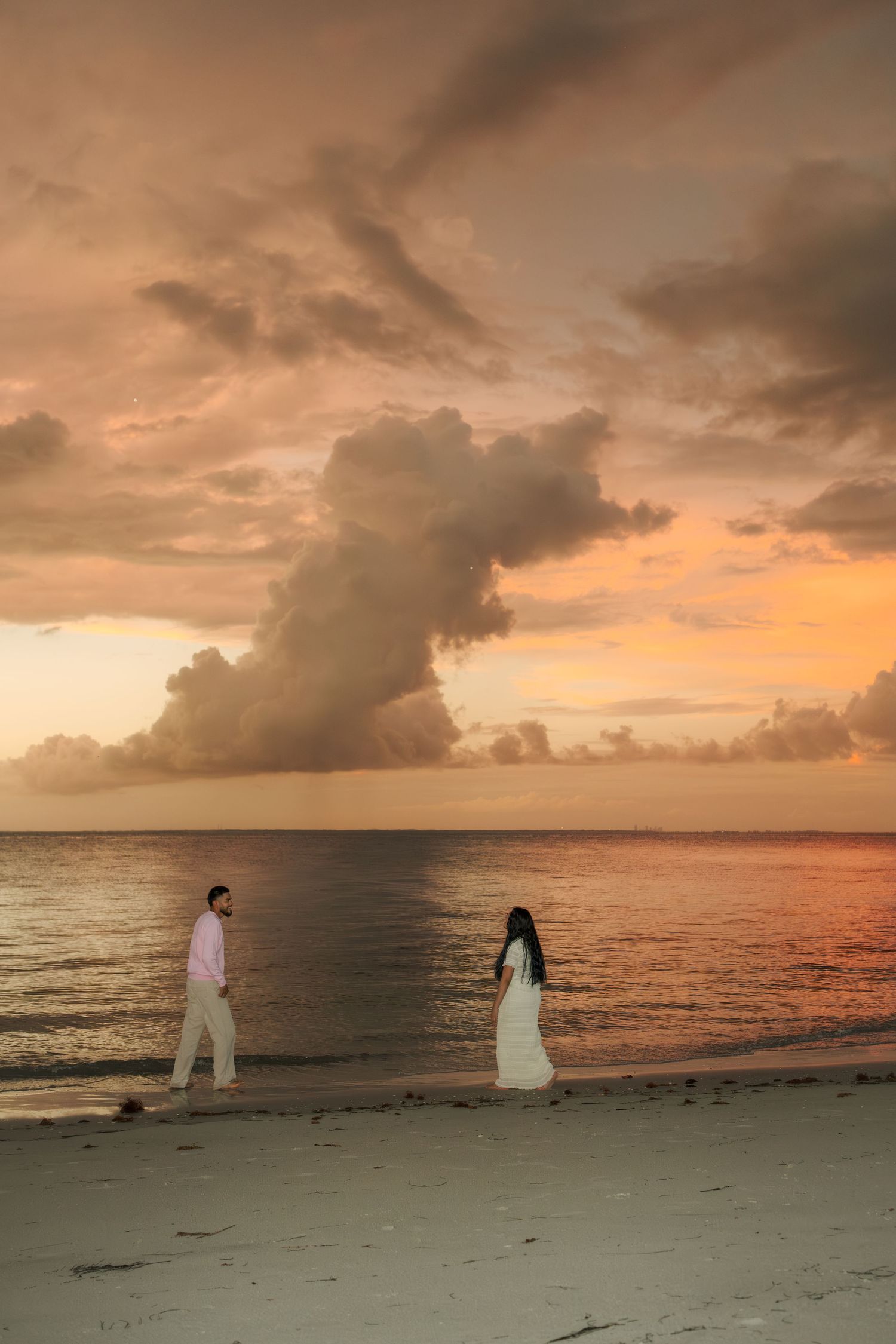 Couple walking on a sandy beach at sunset with dramatic orange clouds over the ocean.
