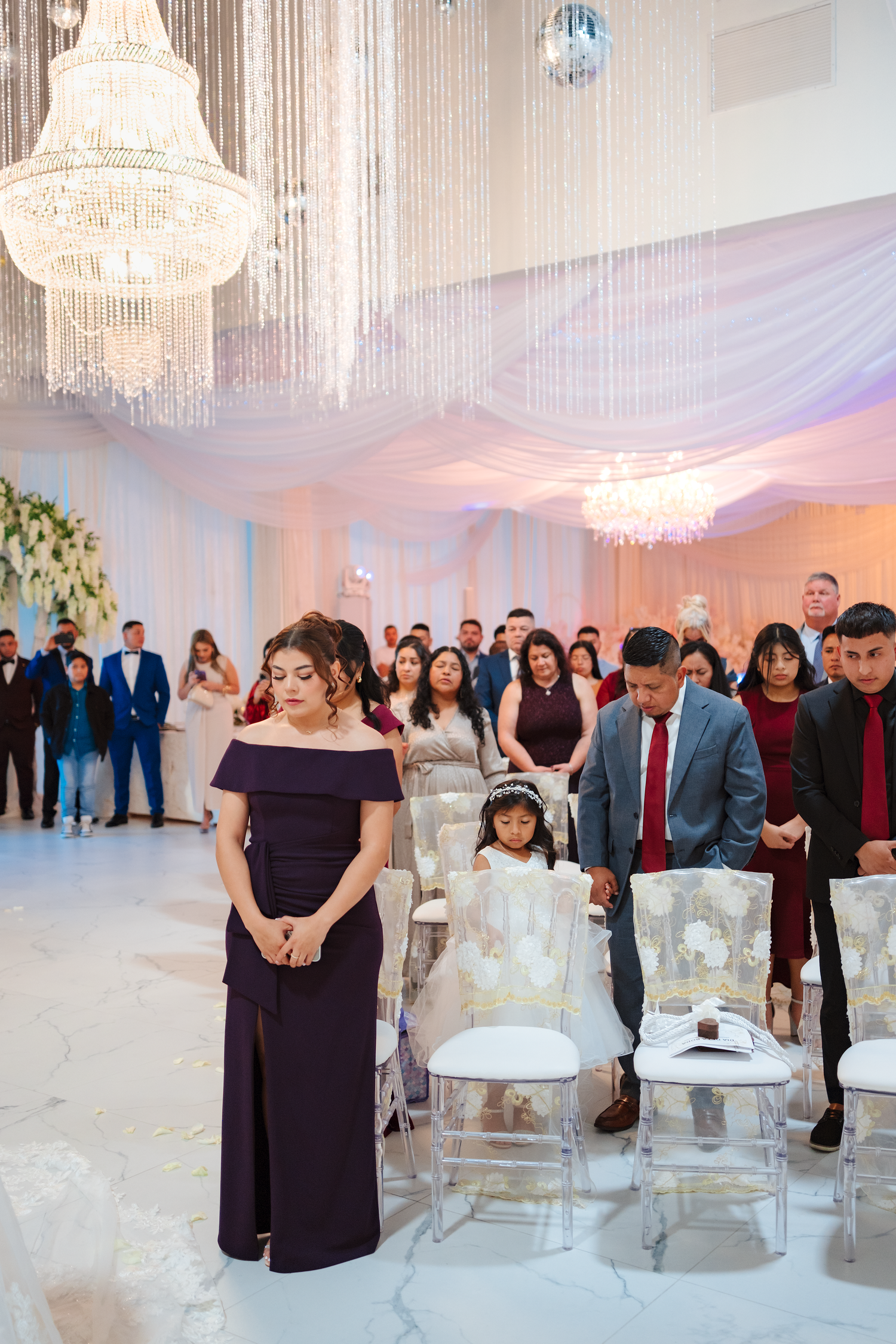 Elegant wedding ceremony guests standing in prayer inside a grand ballroom with crystal chandeliers.