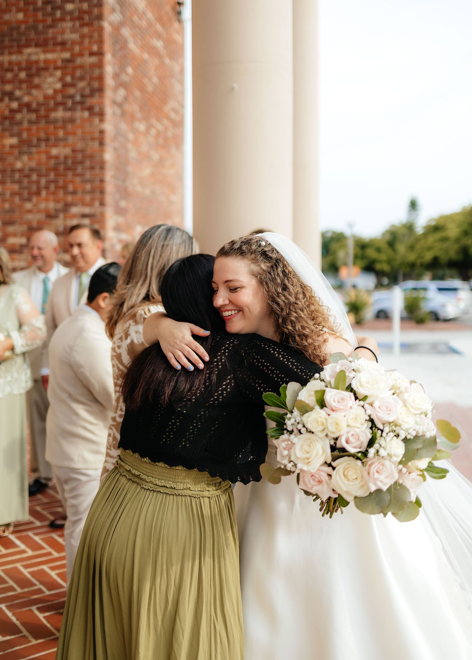 Joyful bride hugging guest outside brick church while holding white rose bouquet.