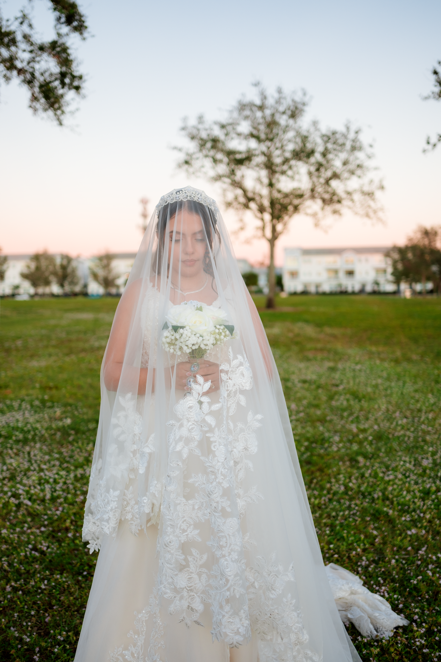 Elegant bride in lace wedding gown and cathedral veil holds white bouquet at twilight outdoors.