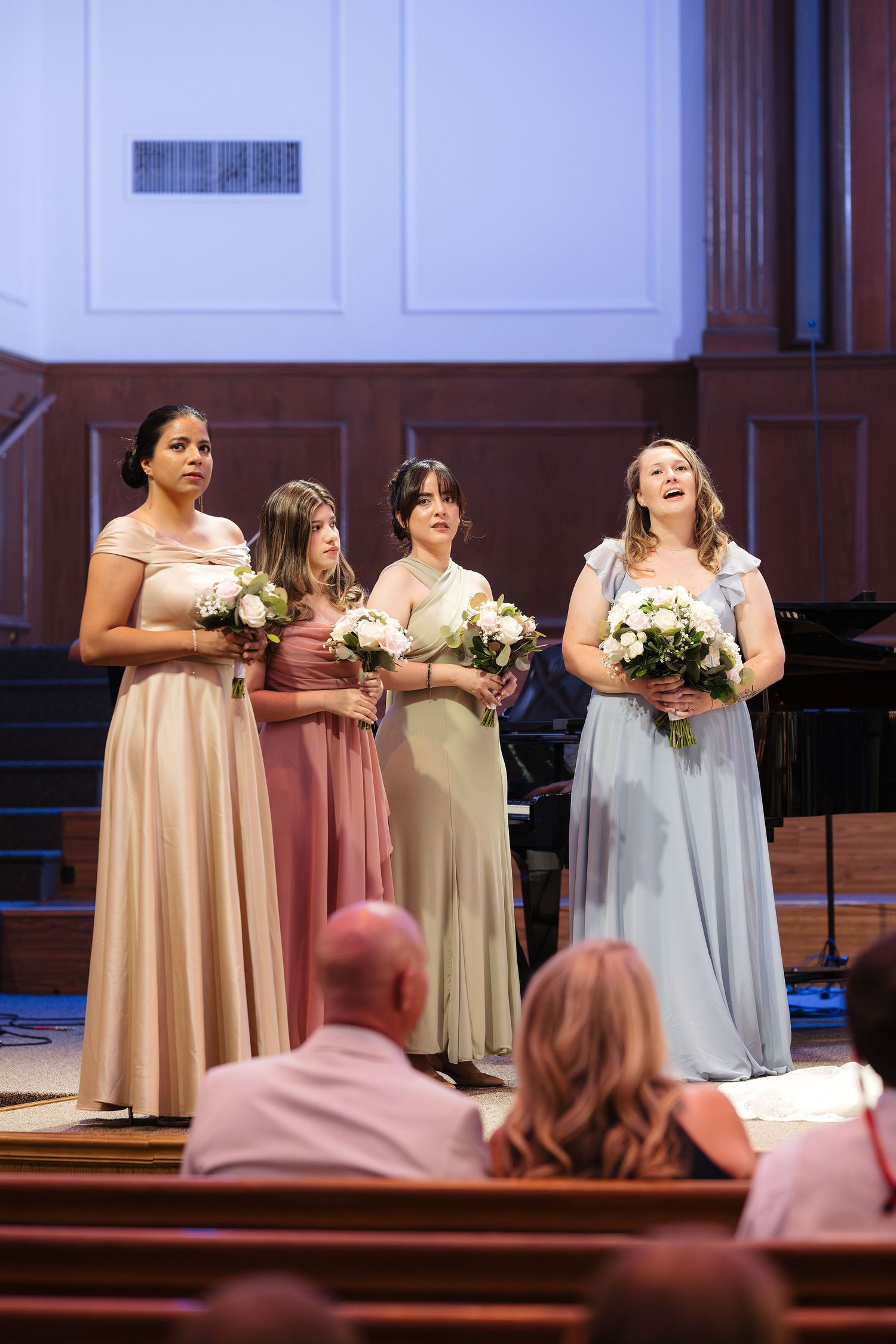 Four bridesmaids in mismatched pastel gowns holding white bouquets stand at the altar during a wedding ceremony.