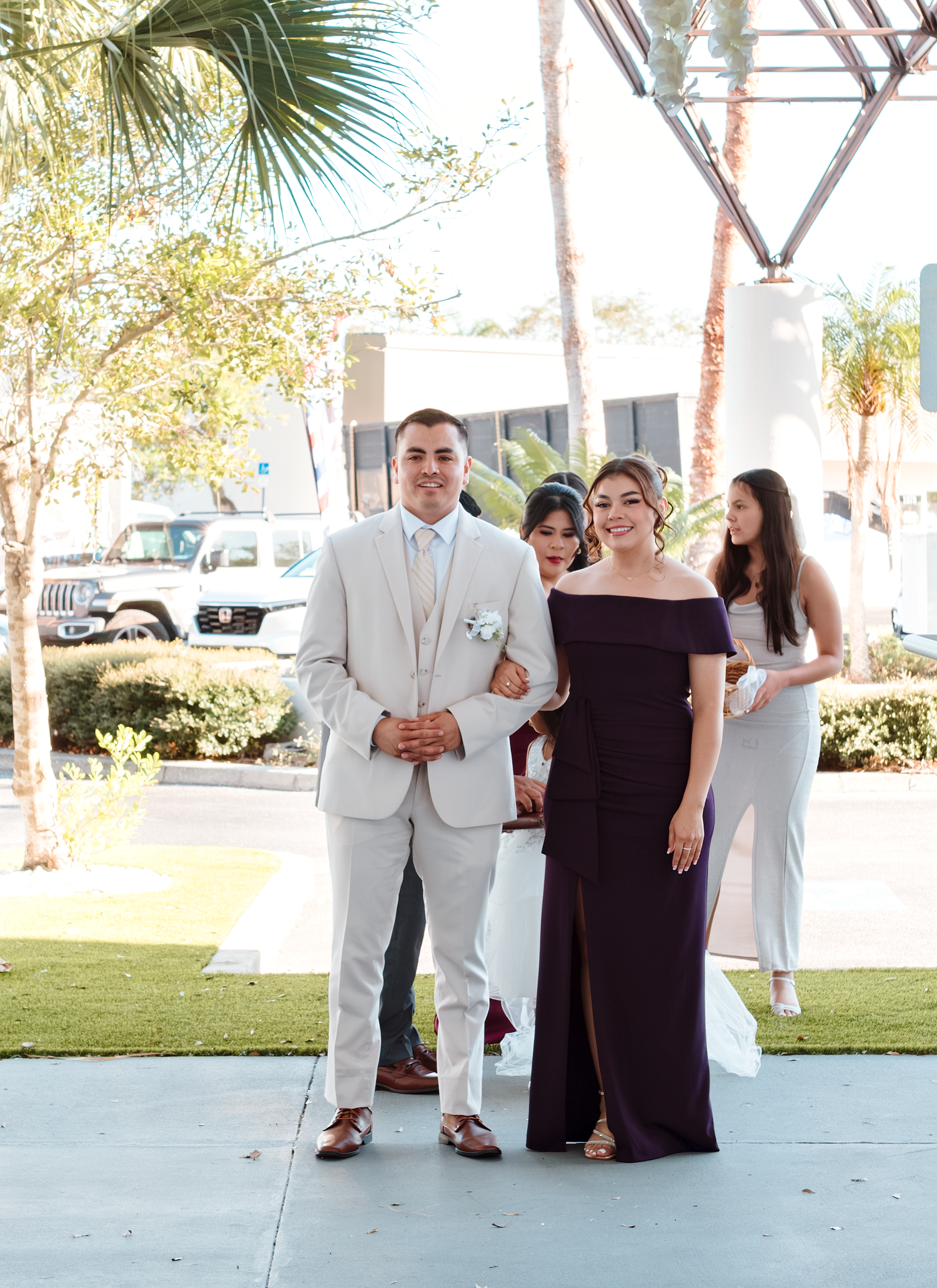 A groom in a light gray suit walks with a bridesmaid in a dark purple off-shoulder gown at an outdoor wedding.