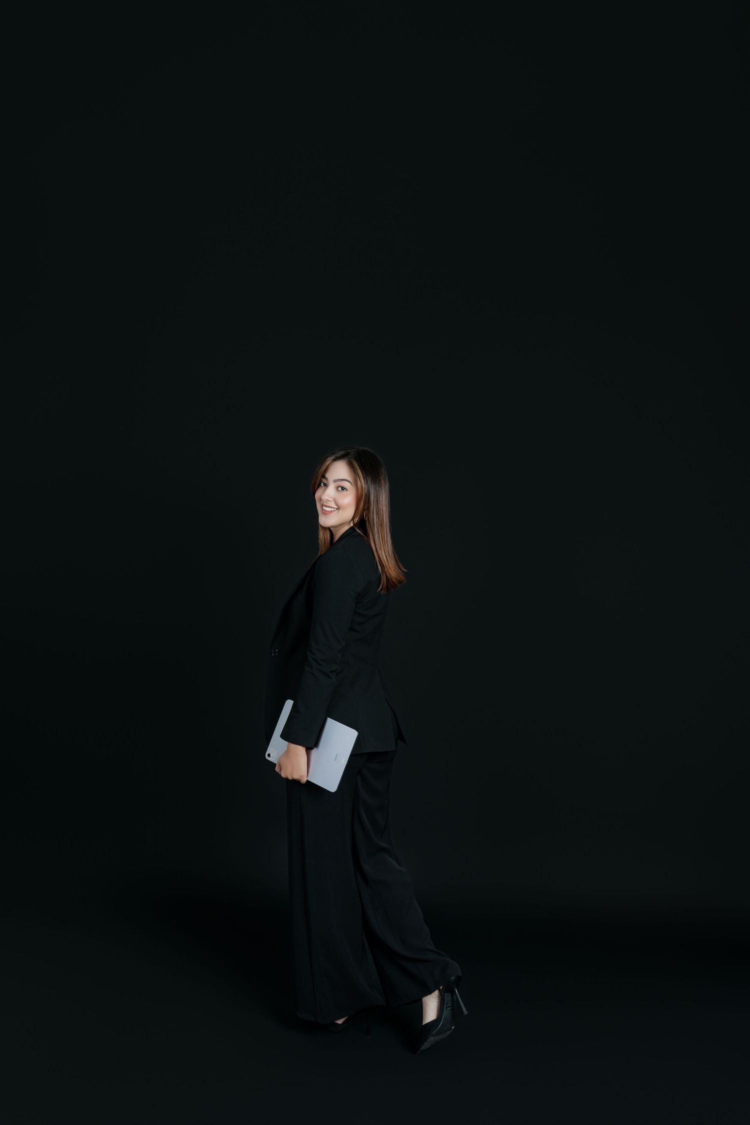 Confident businesswoman in black suit holding laptop turning to smile in dark studio.