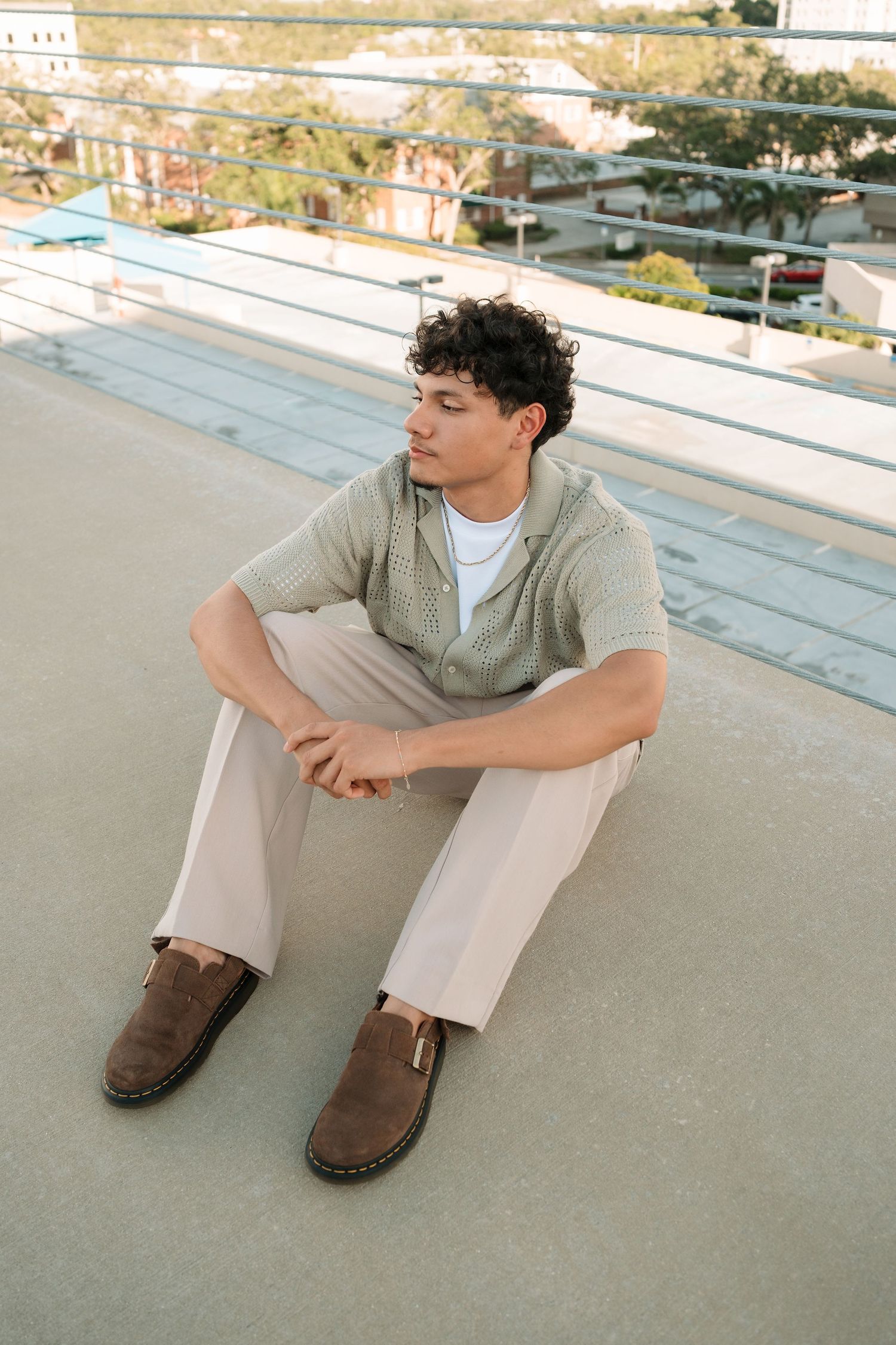 Young man in sage knit shirt and cream trousers sitting on rooftop, wearing brown loafers, looking away thoughtfully.