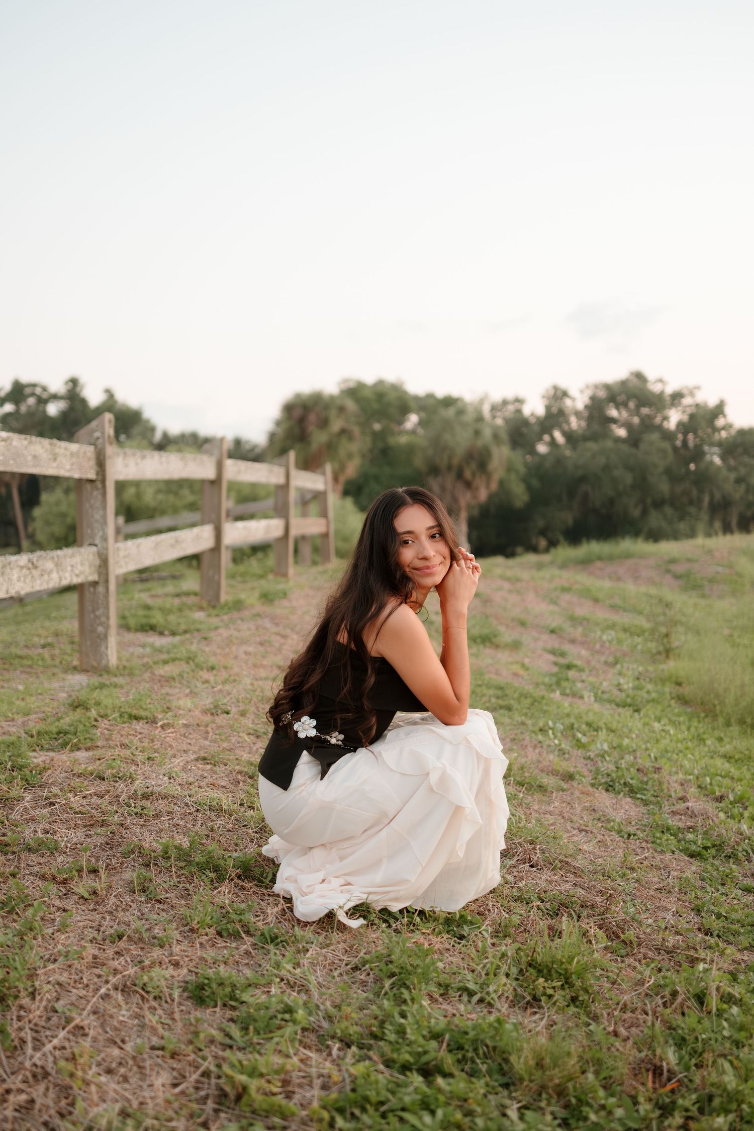 Brunette bride crouching in white skirt and black corset top near wooden fence in a pastoral setting.