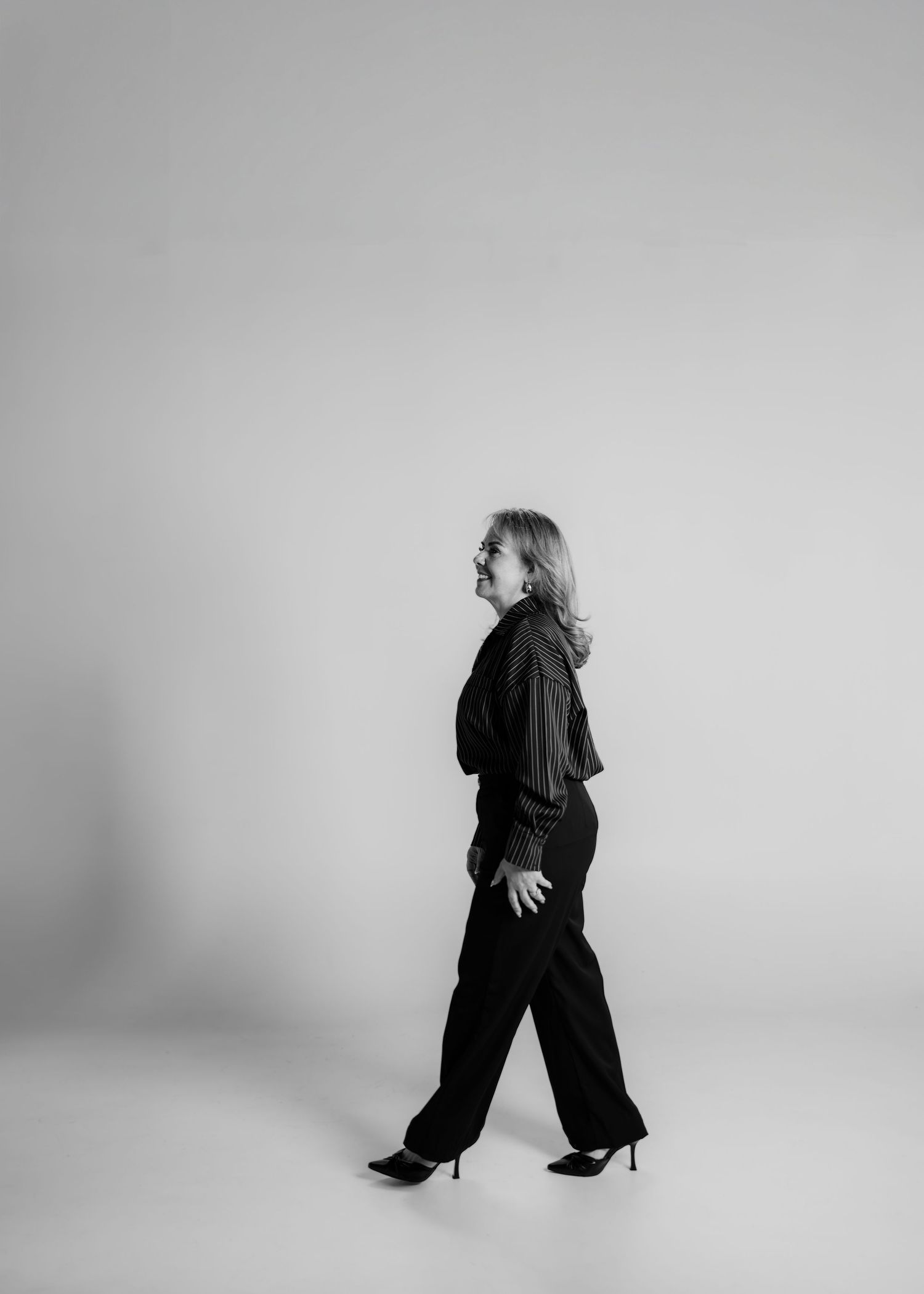 Black and white photo of a blonde woman walking in black wide-leg pants, heels, and a dark blouse against a gray backdrop.