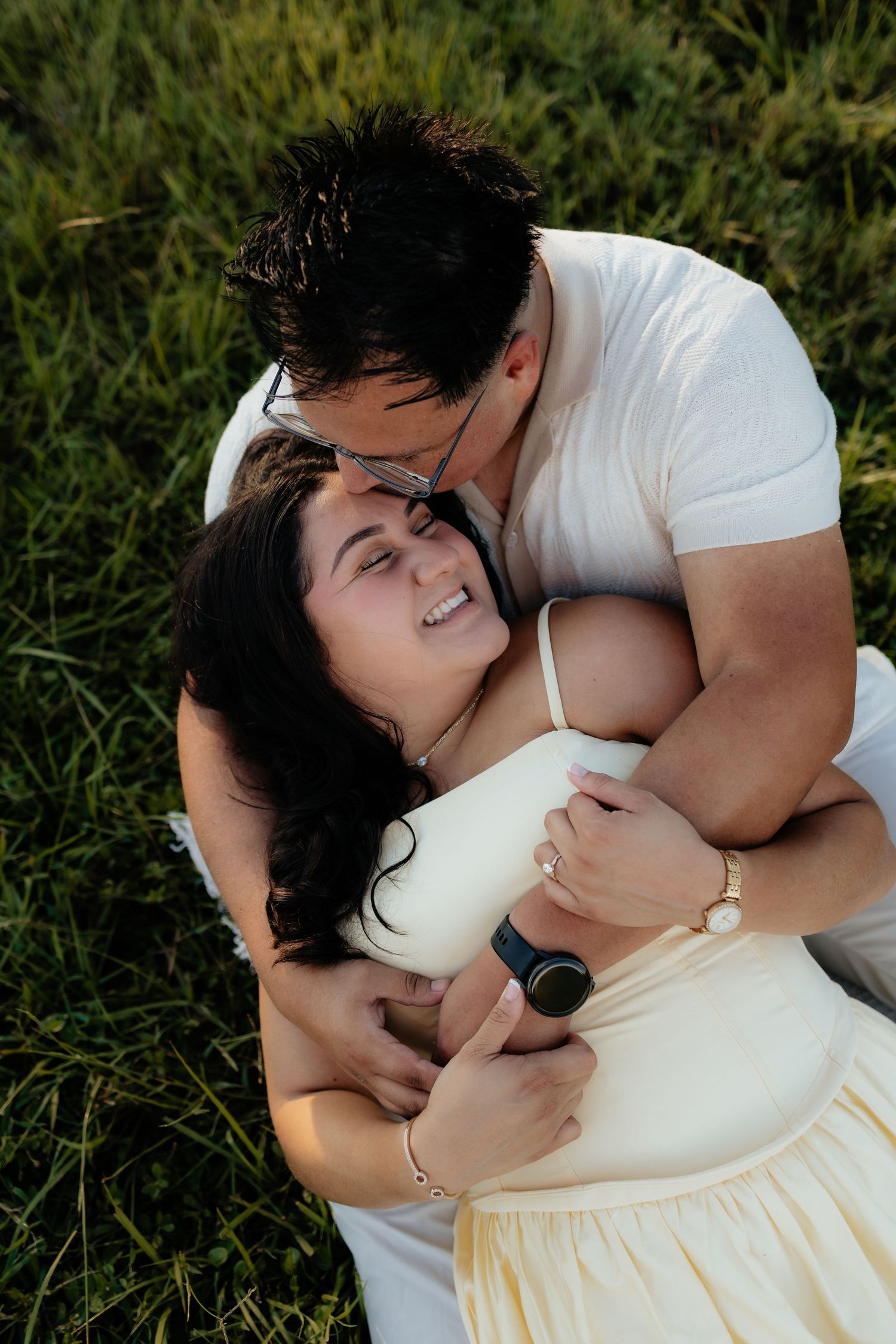 Couple sharing a tender moment on green grass, man kissing womans forehead while she laughs joyfully.