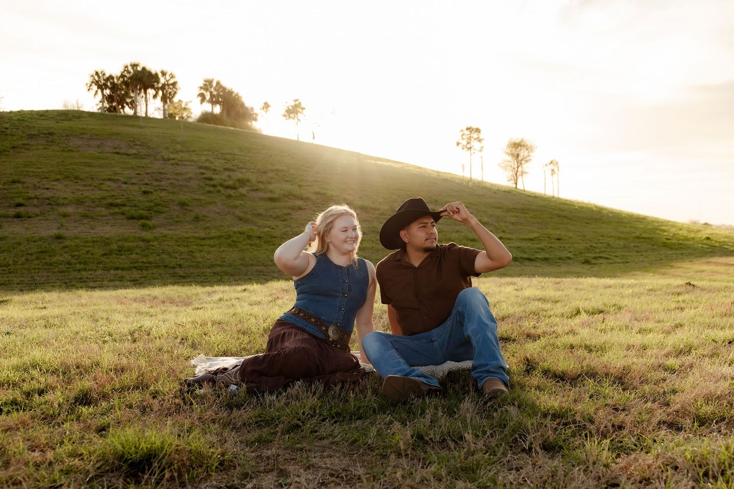Couple sitting on green hillside at golden hour, woman in blue dress and man in cowboy hat enjoying sunset.