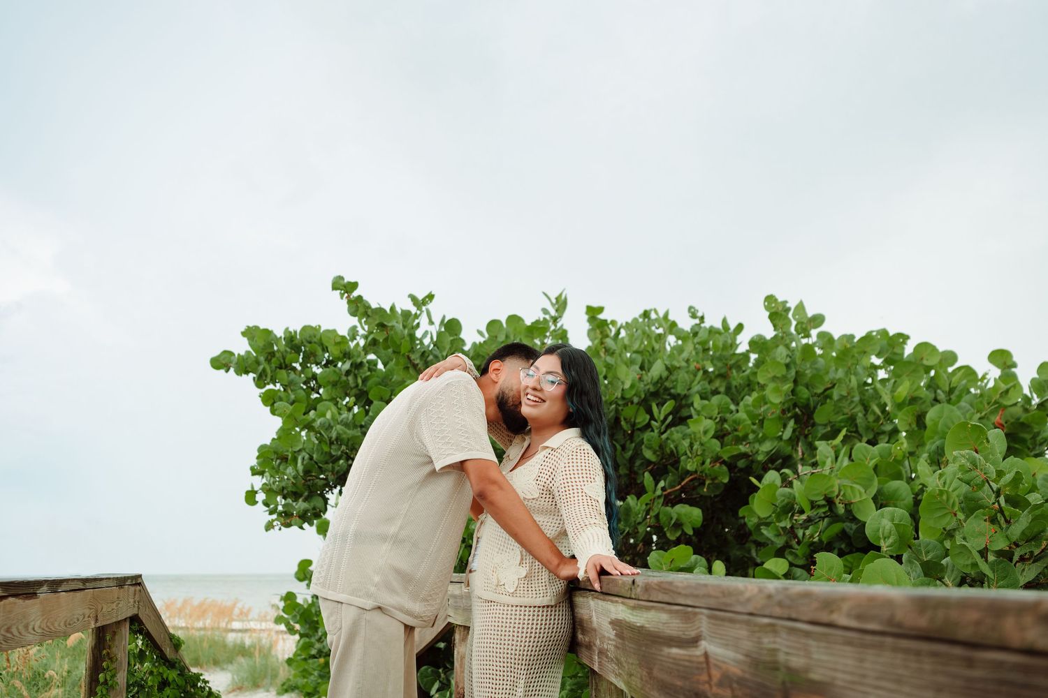 Couple sharing a romantic kiss on a wooden boardwalk surrounded by lush green tropical plants near the beach.