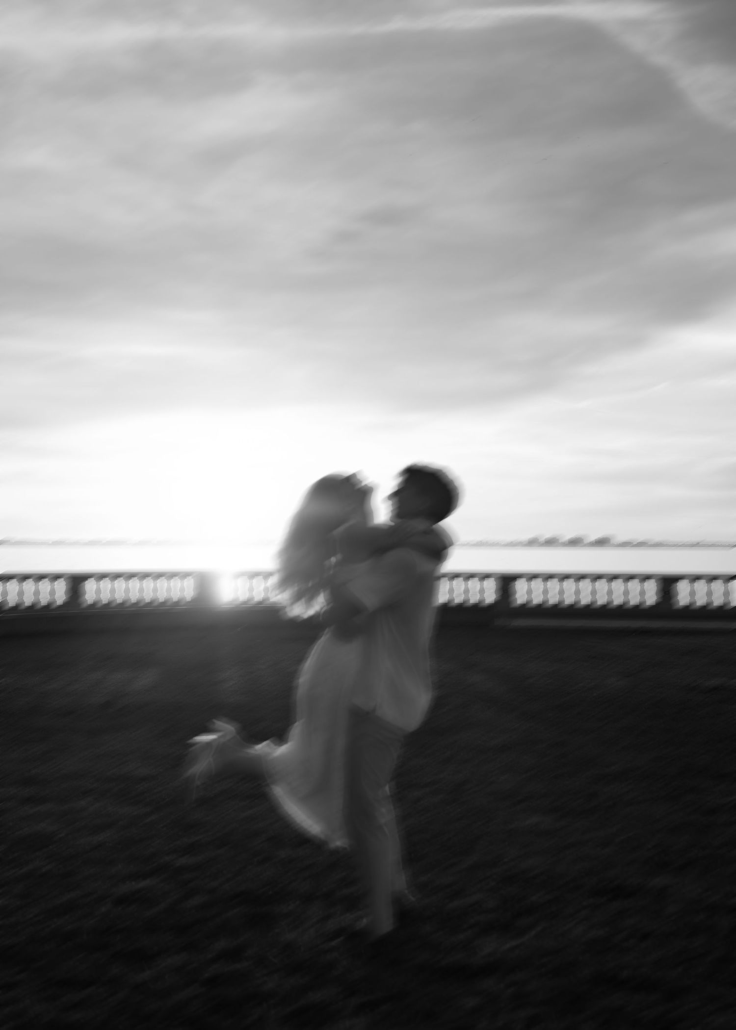Black and white photo of a couple embracing at sunset, silhouetted against a glowing horizon near a balustrade.