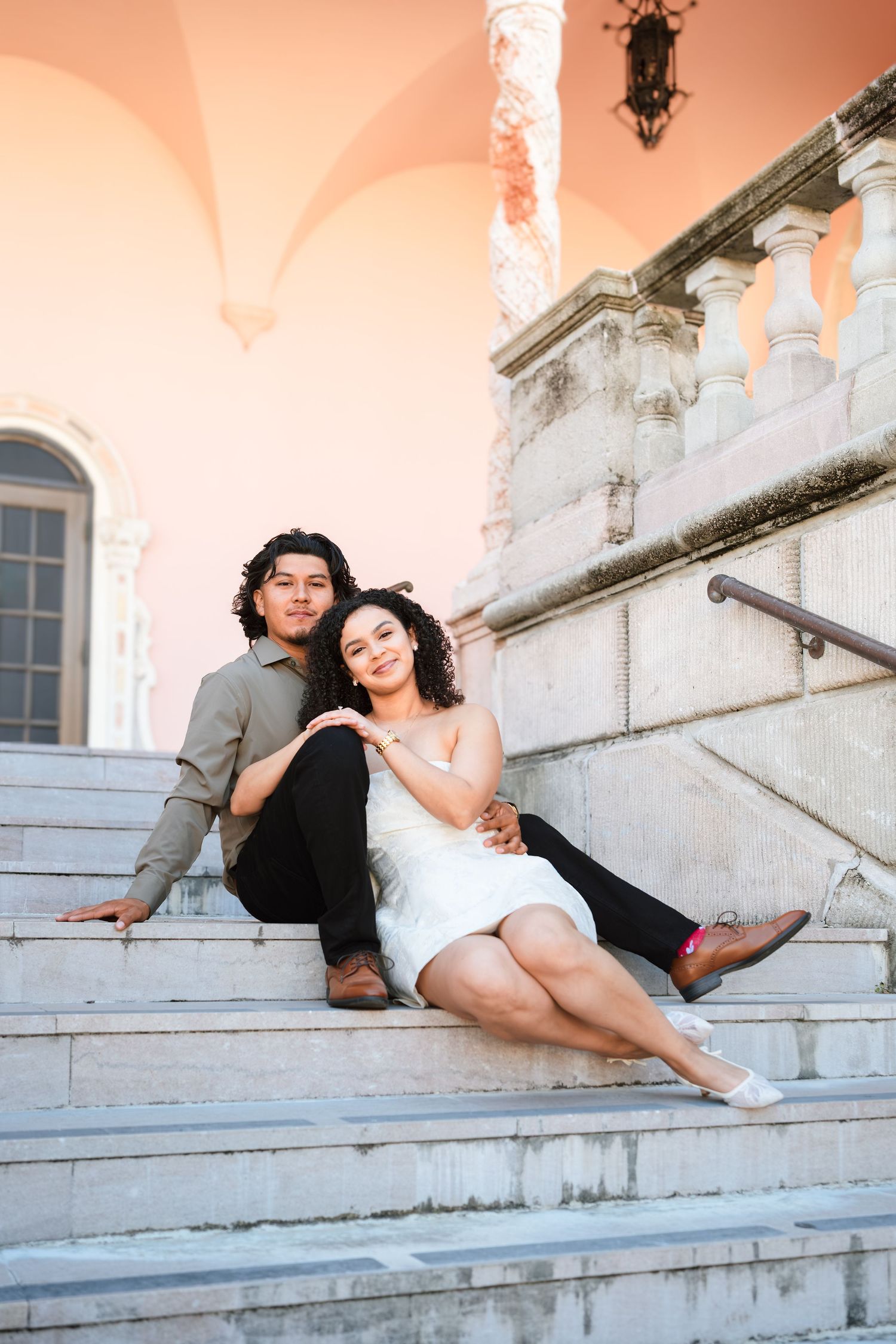 A young couple sits together on marble steps of an elegant building, the woman in a white dress leaning against the man.