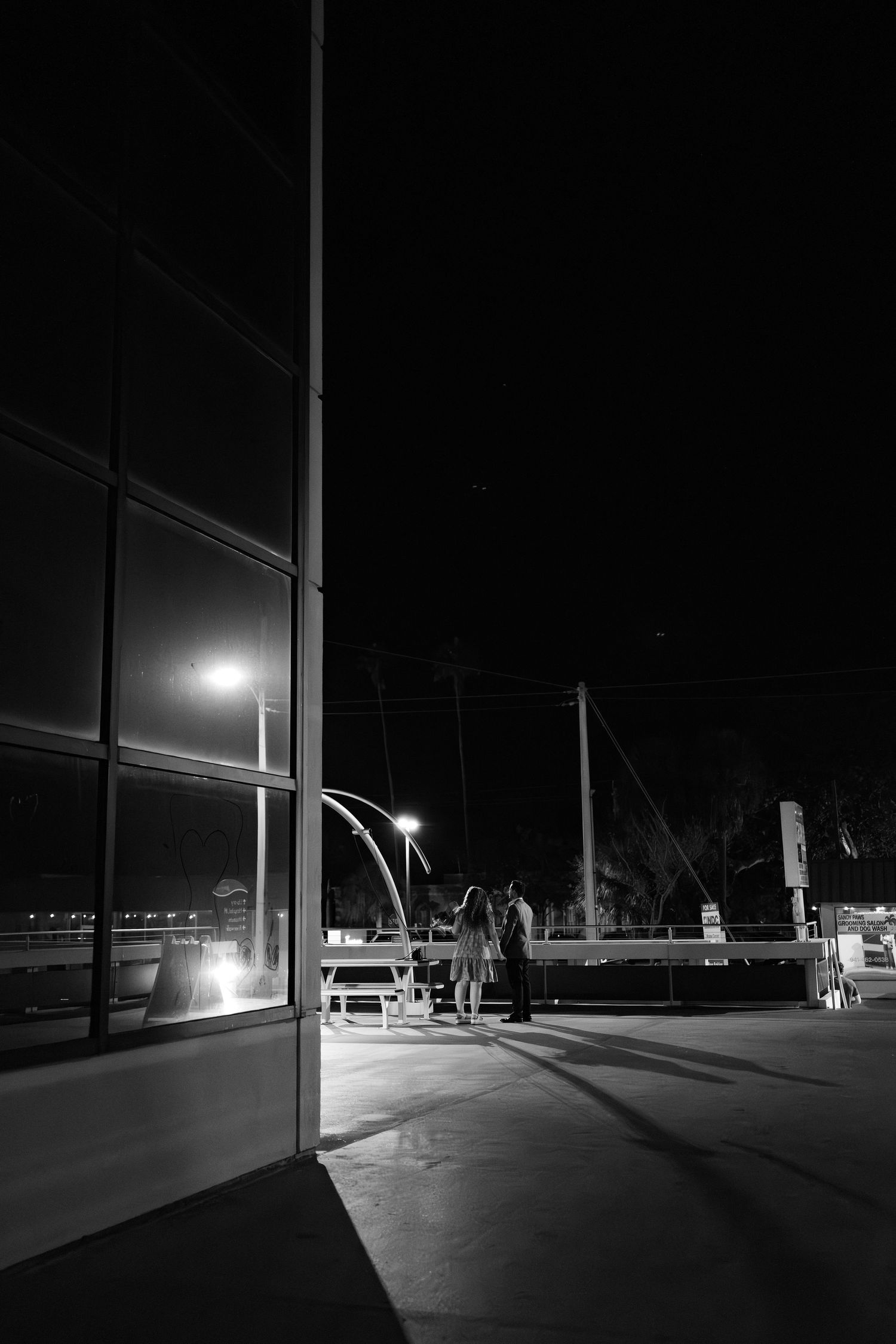 Black and white night photo of two people standing outside near street lights and a glass building.