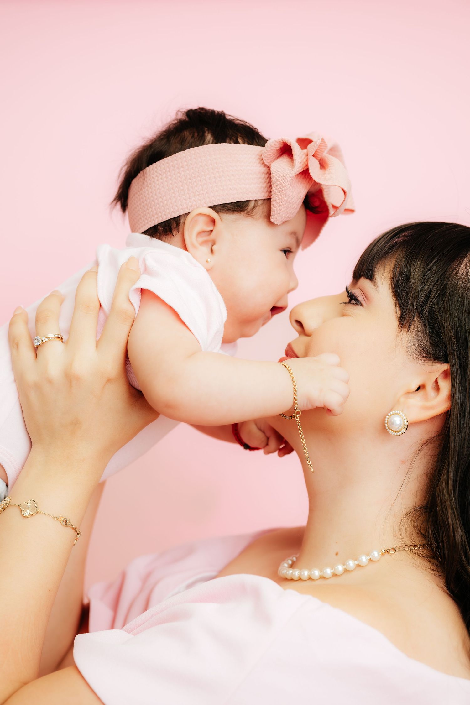 A mother holds her baby girl close, both wearing pink accessories against a soft pink background.