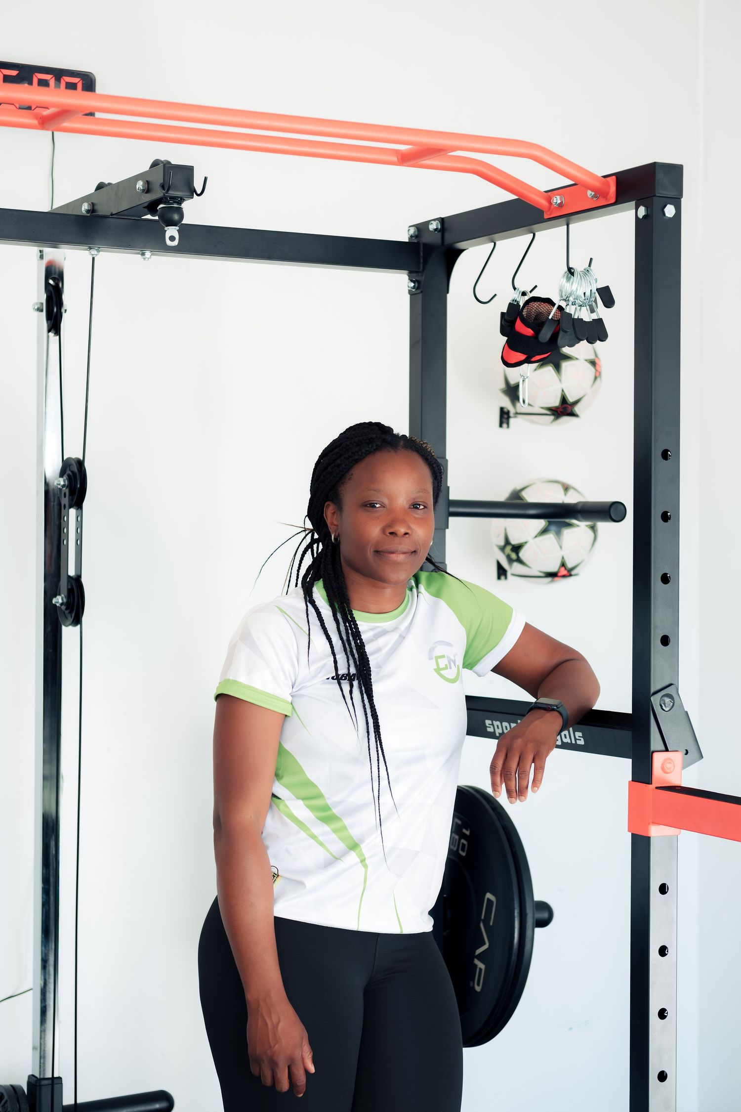Woman in white and green athletic shirt posing confidently in a gym next to a power rack with weight plates.