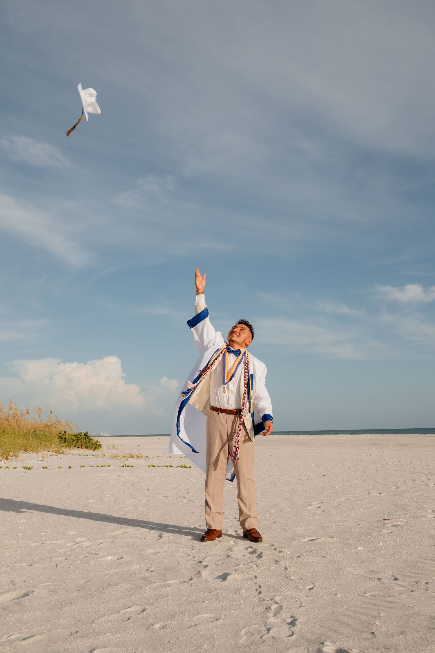 Man in white robe tossing graduation cap on a sunny beach with blue sky.