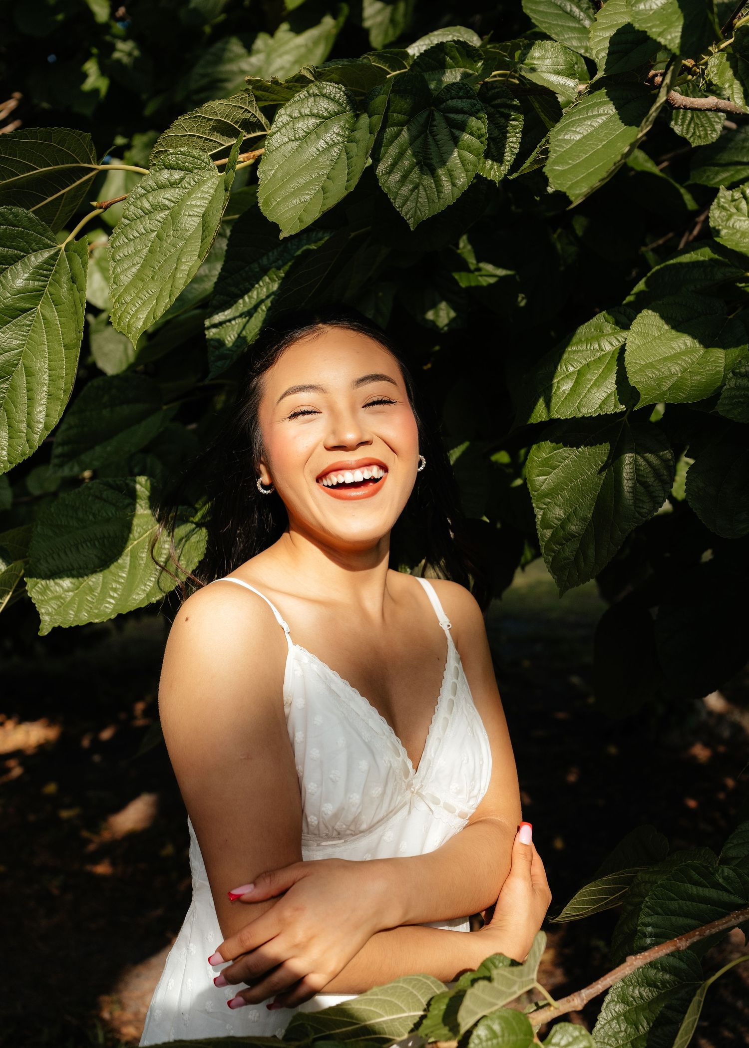 A young woman in a white dress laughs joyfully surrounded by lush green leaves in sunlight.