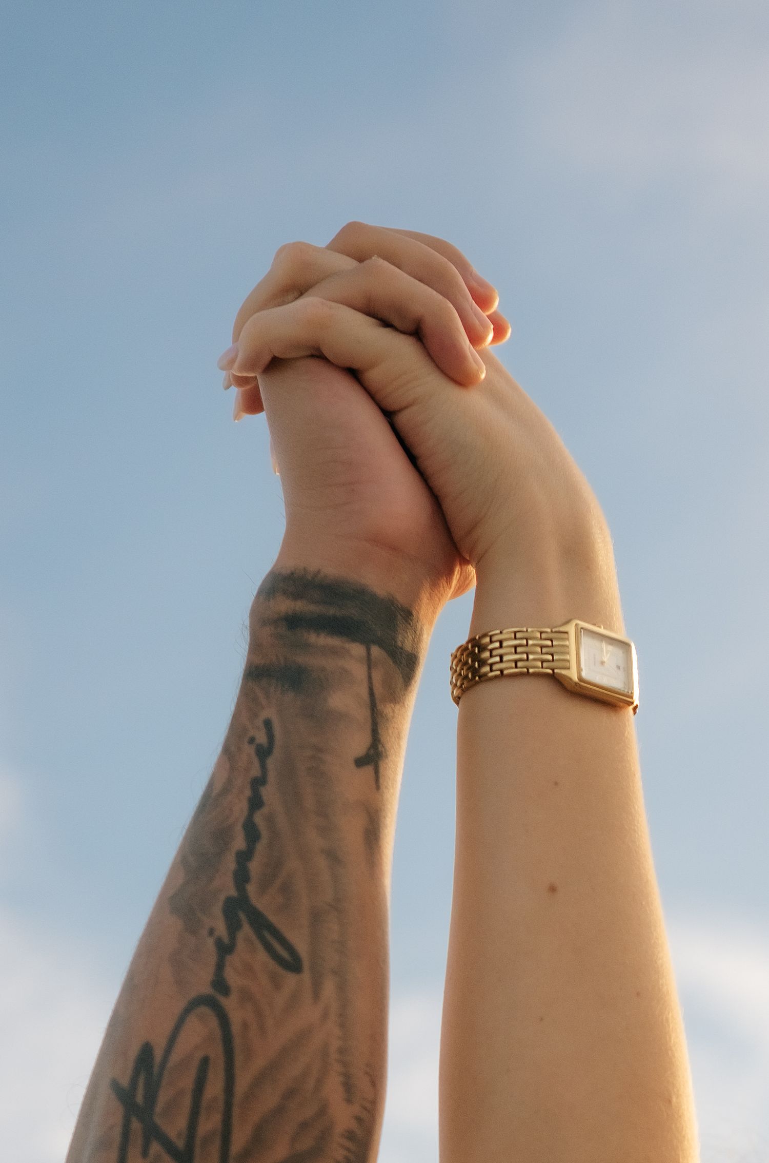 Two hands clasped together raised against blue sky, one with tattoos and one wearing a gold watch.