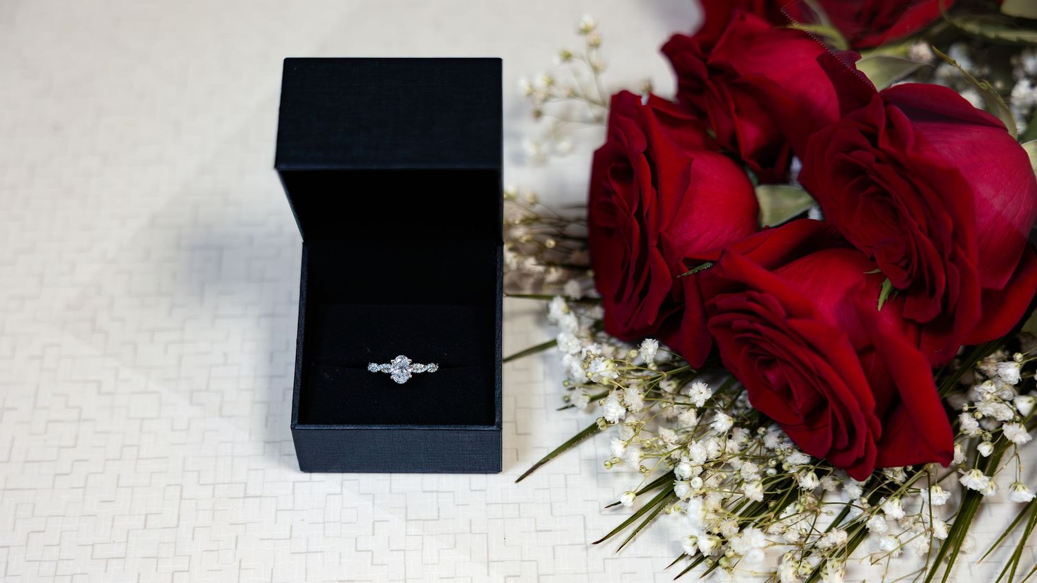 Diamond engagement ring in black box beside red roses and baby's breath on white surface.