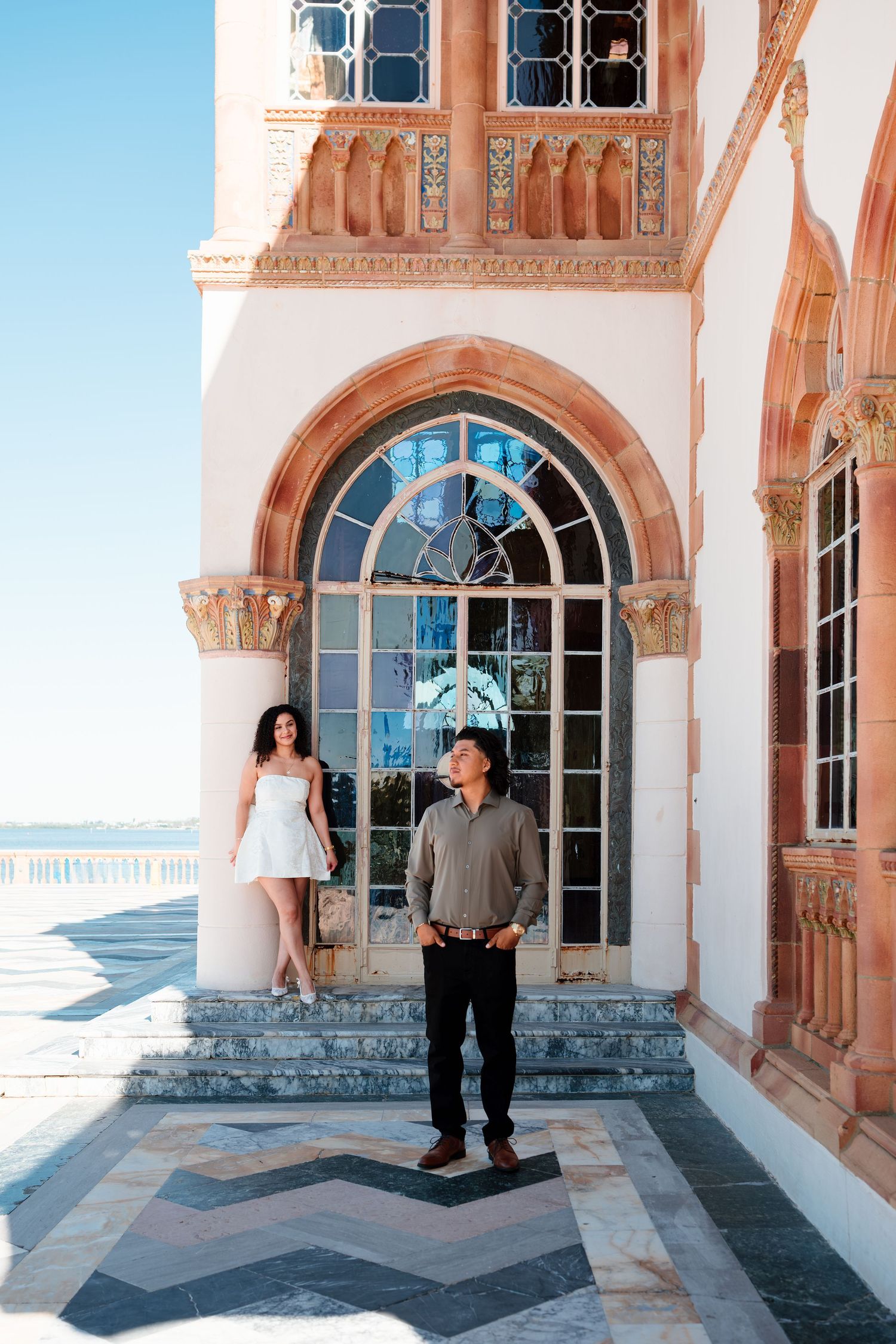 Couple posing at ornate Mediterranean-style building with arched windows and ocean views on sunny day.