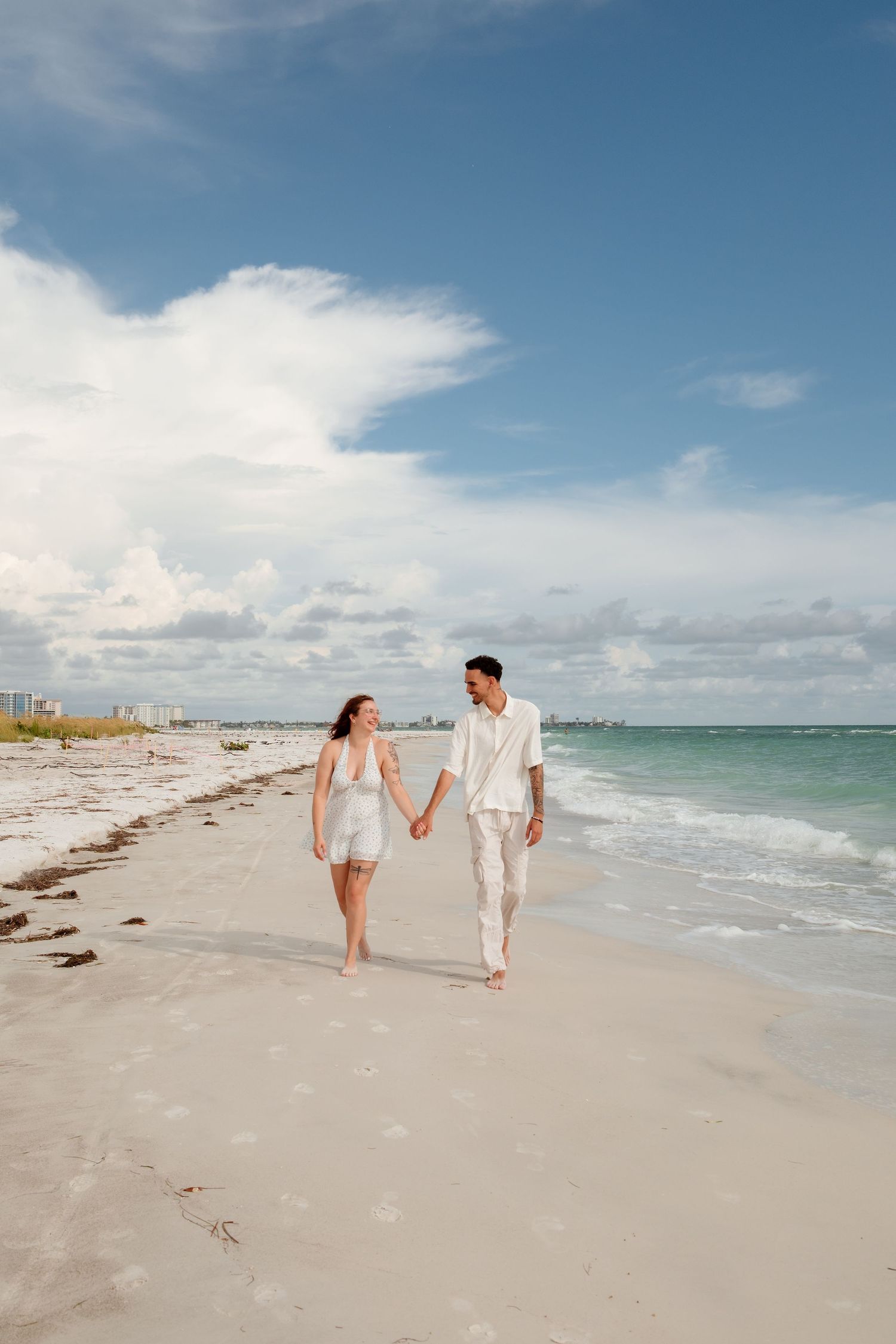 Couple holding hands walking on a sunny white sand beach with turquoise ocean waves and blue sky.