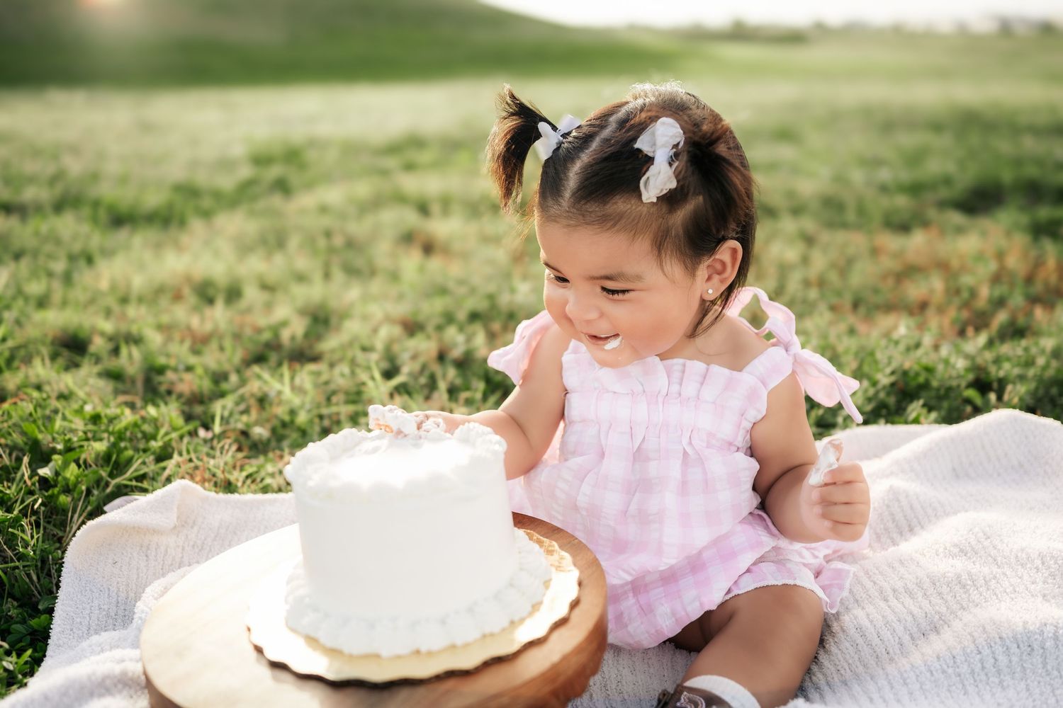 A series of photos showing a cake smash photo session with a baby in a pink dress on a white blanket outdoors.