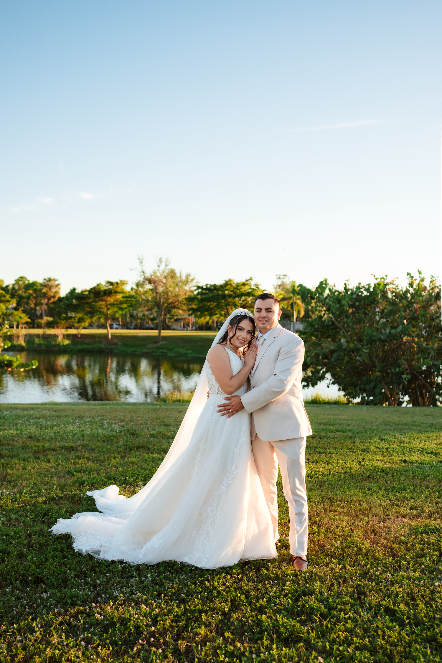 Bride and groom embracing outdoors by a serene lake at golden hour in wedding attire.