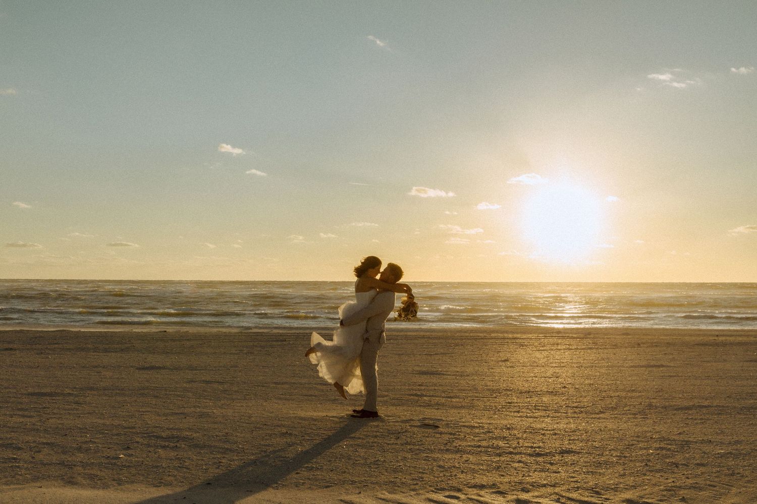 Couple sharing a romantic kiss at sunset on a beach, groom lifting bride in wedding attire with golden light.