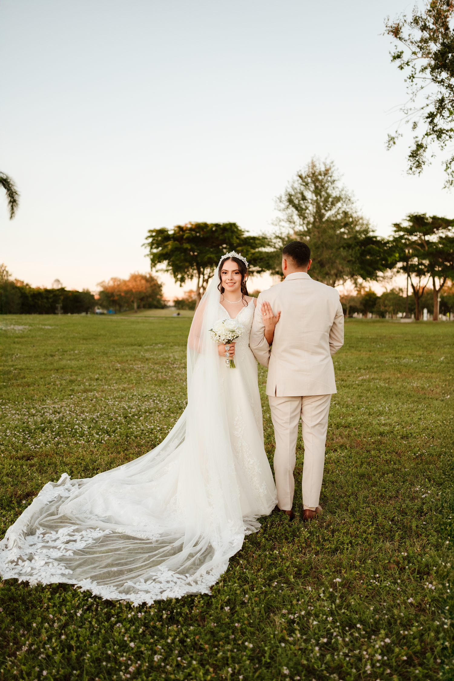 Bride in a long lace veil gown holds bouquet while groom in beige suit stands in a sunlit green outdoor setting.