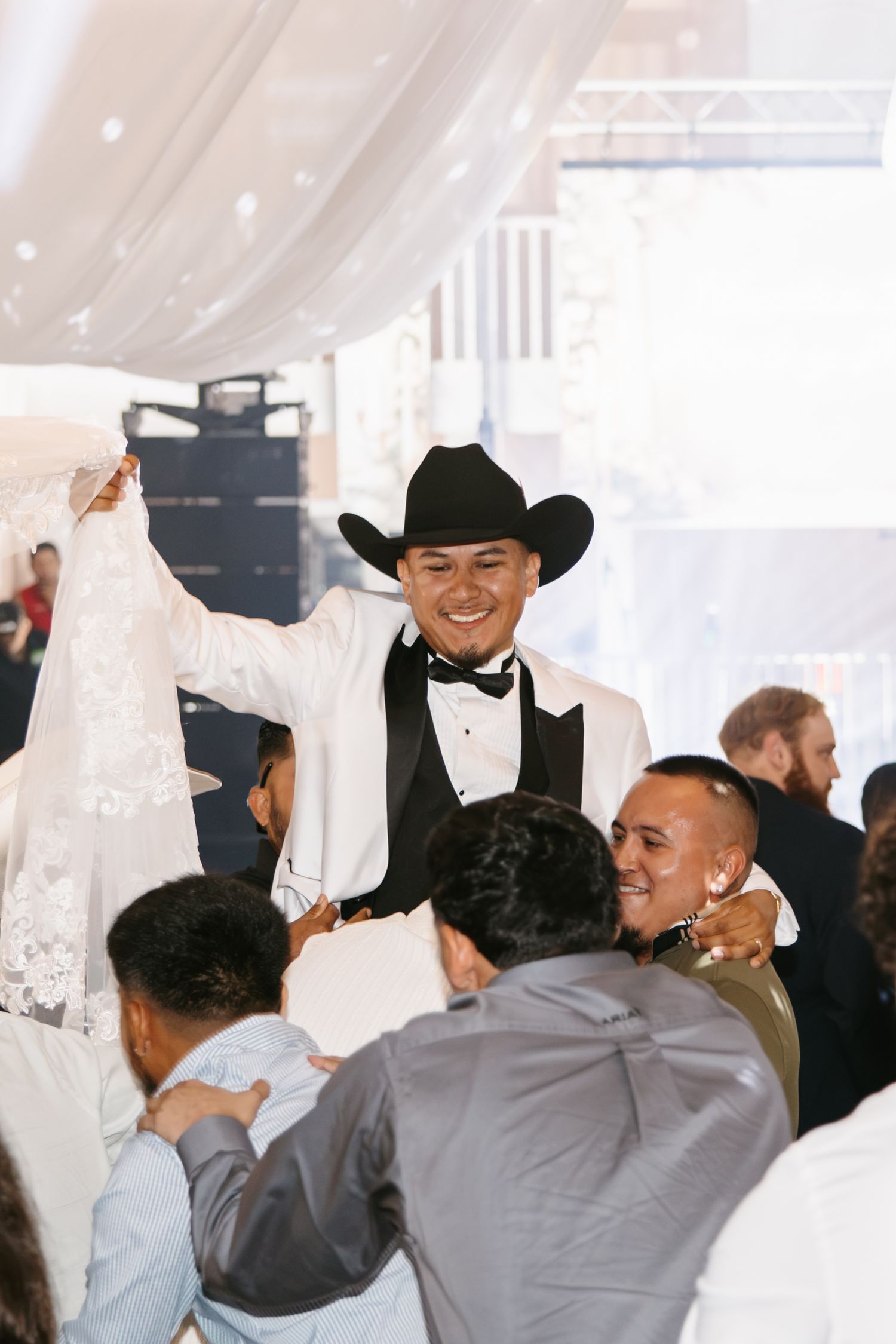 Groom in white tuxedo and black cowboy hat smiles at wedding reception surrounded by celebrating guests.