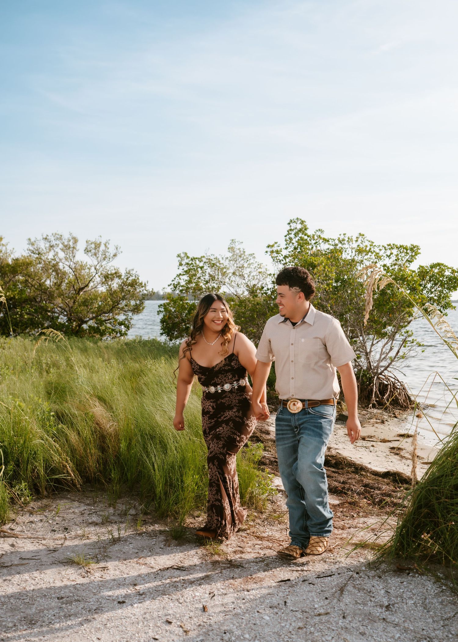 Couple walking hand in hand on sandy path surrounded by tall green grass and water under a sunny blue sky.