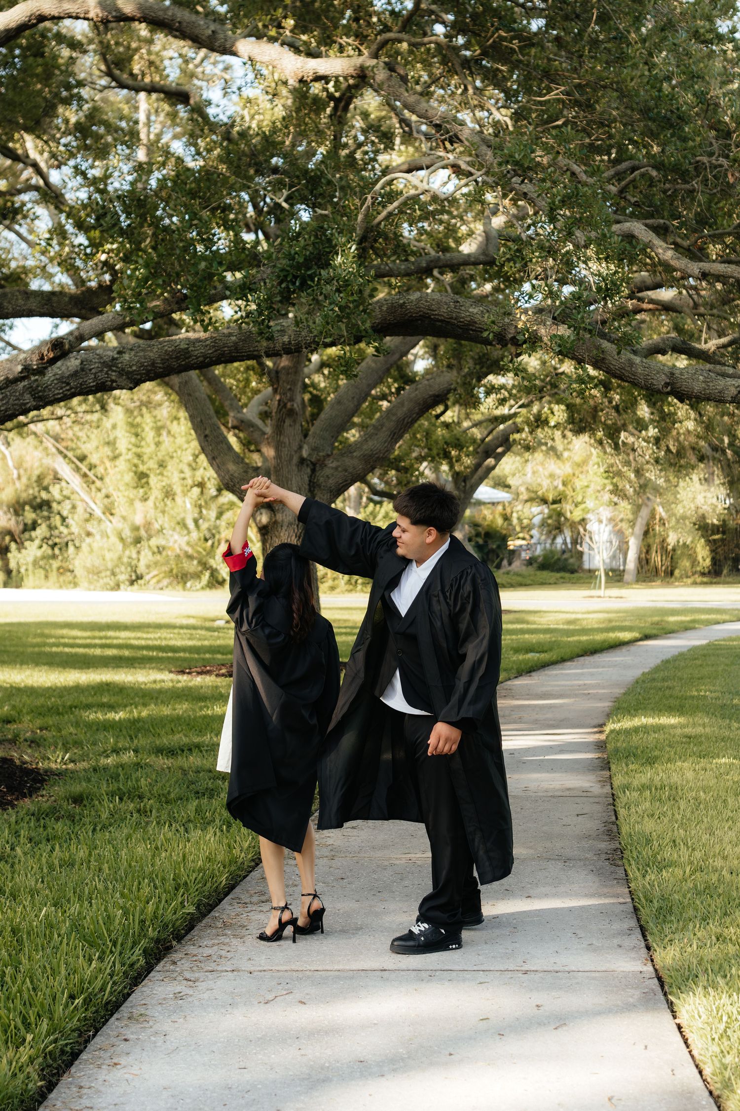 Two graduates in black gowns dance on a sunny campus walkway surrounded by large oak trees and green lawns.