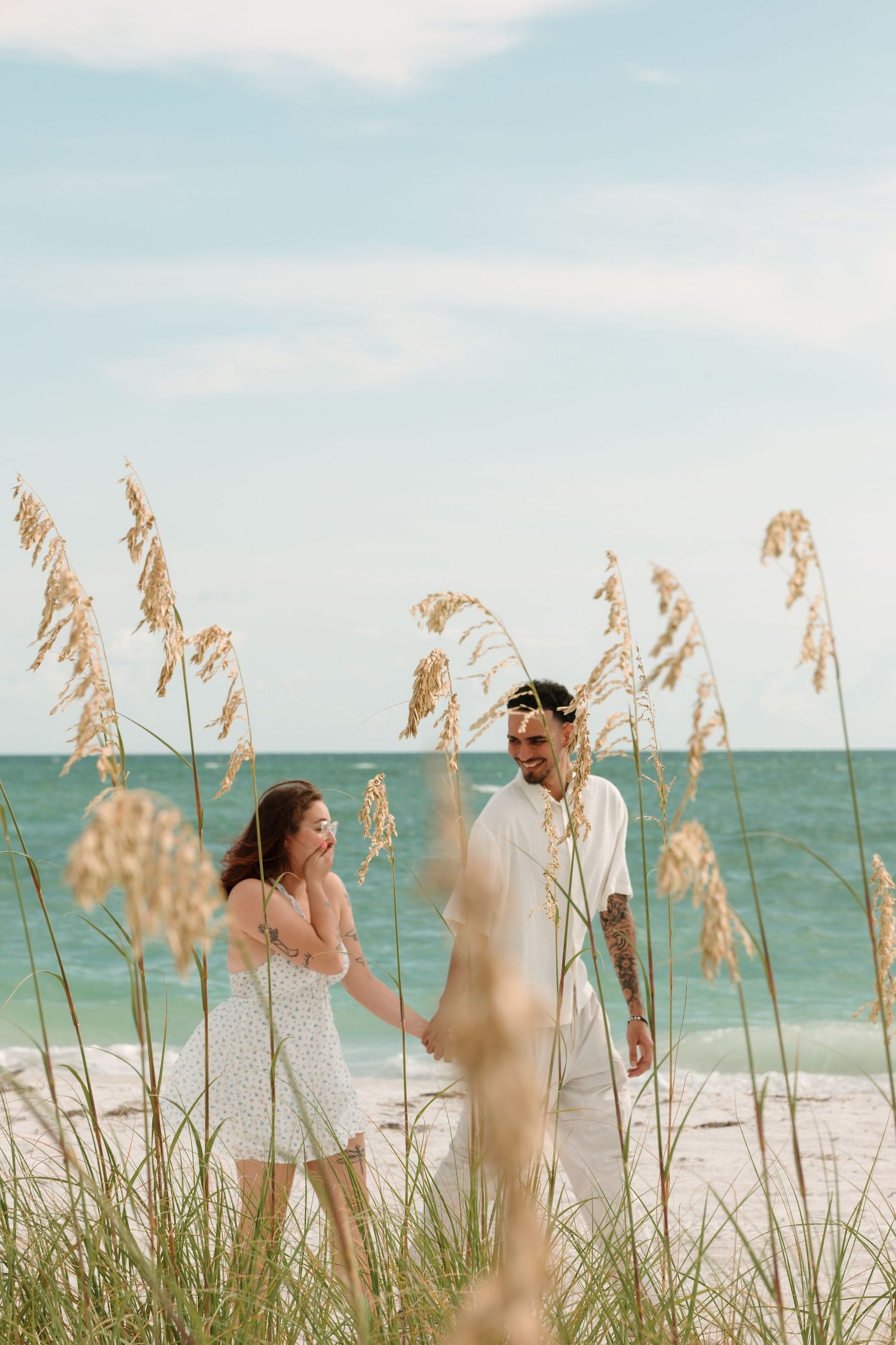Couple holding hands on a sandy beach surrounded by sea oats with turquoise ocean waves in the background.