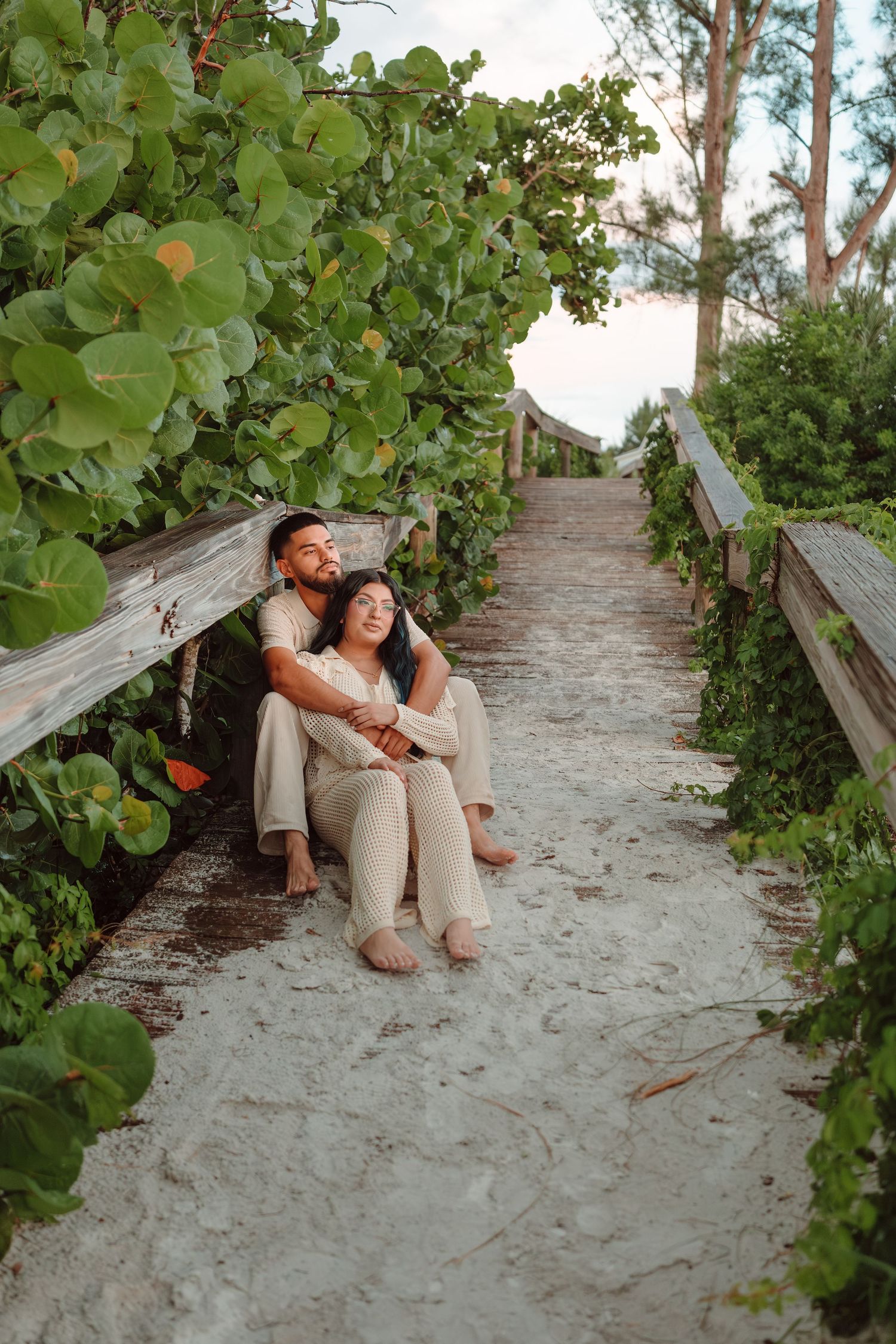 A couple sits together on a sandy beach boardwalk path, surrounded by lush green tropical foliage at sunset.
