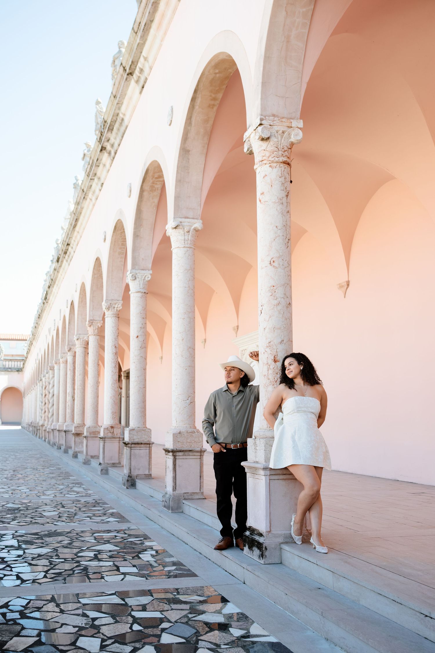 Elegant couple standing among arched pink colonnade, man in cowboy hat, woman in white strapless dress.