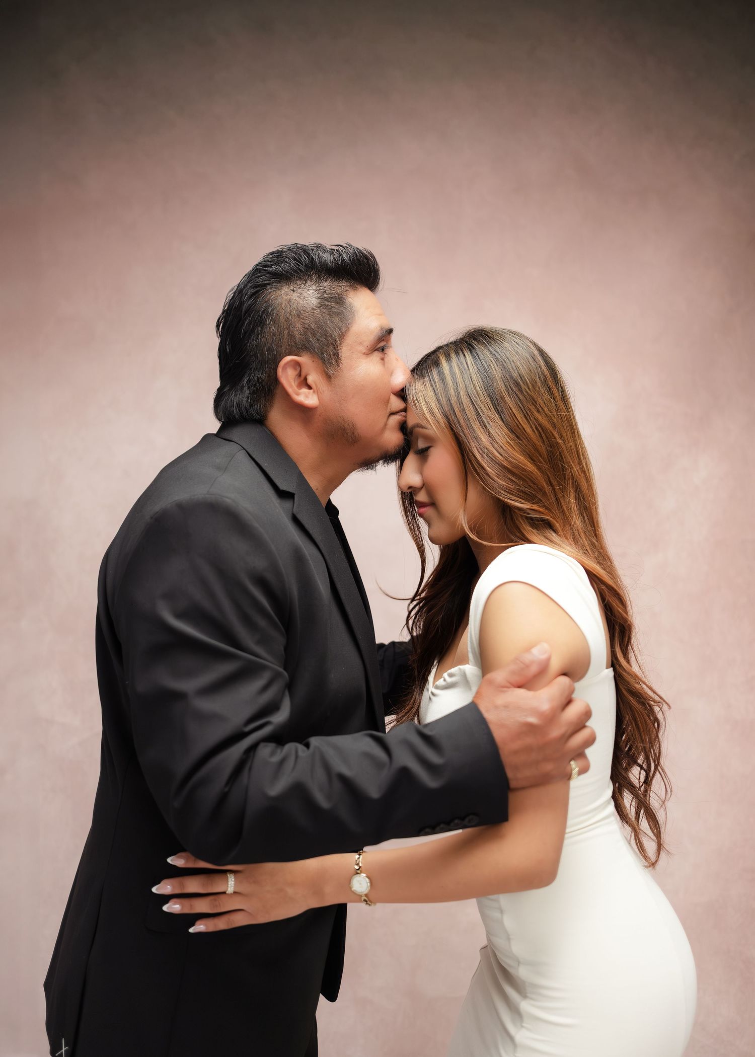 A man in a black suit kisses a woman in a white dress on the forehead during a romantic studio portrait session.